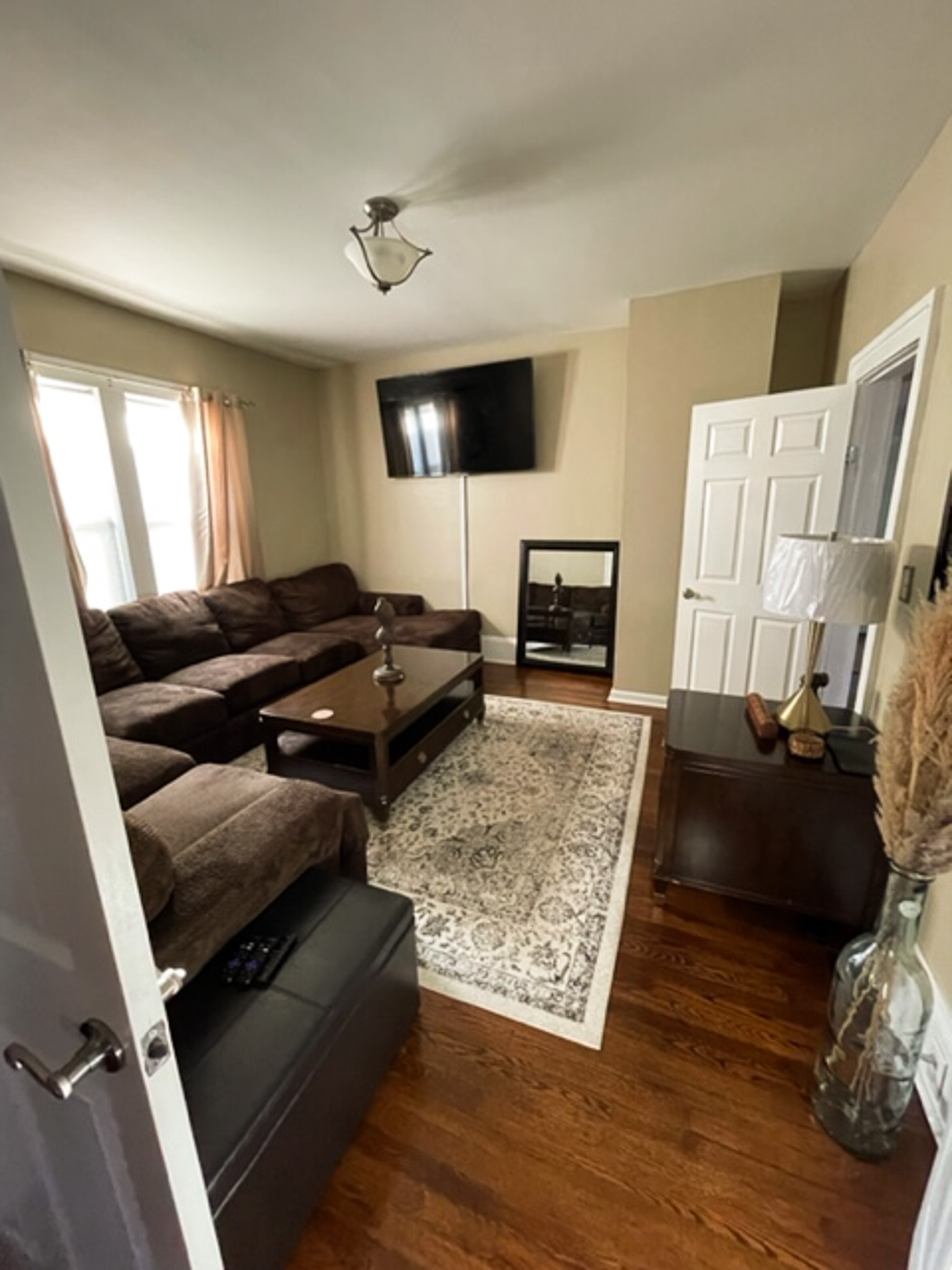 A living room with a large sectional sofa, patterned rug, wall-mounted TV, and wooden floors inside the home of Sponsored Residential Provider Jessica Peregrino in Roanoke, Virginia.