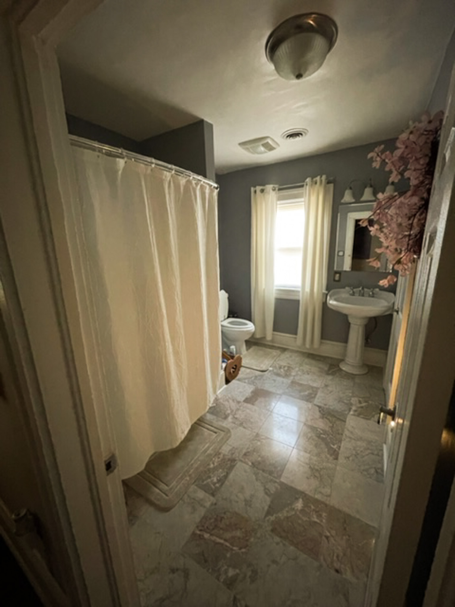 A bathroom with a shower curtain, pedestal sink, toilet, and a window with sheer curtains inside the home of Sponsored Residential Provider Jessica Peregrino in Roanoke, Virginia.