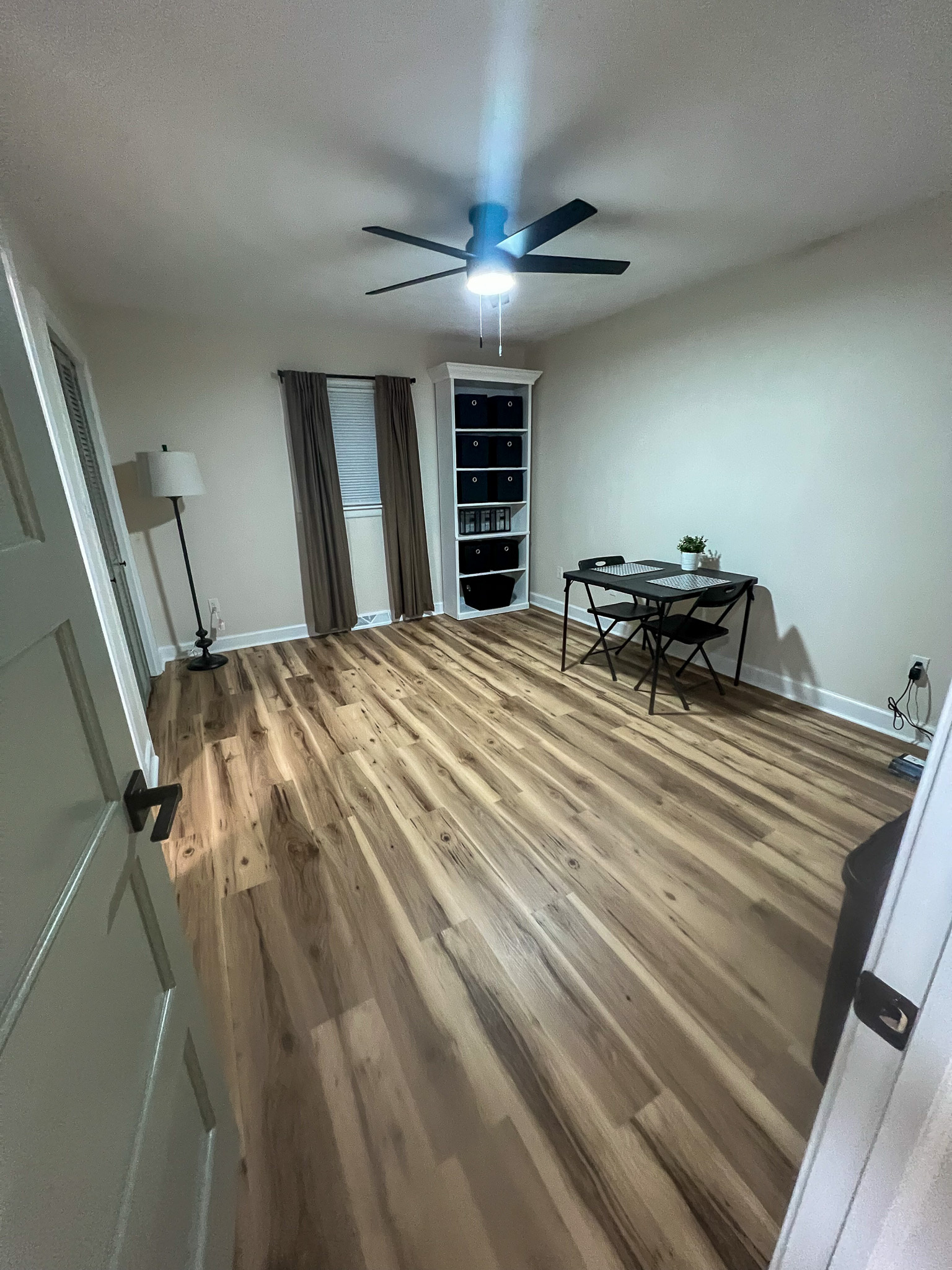 A spacious room with wood floors, a ceiling fan, a small desk and chair, and a window with brown curtains inside the home of sponsored residential provider Regina Hill in Roanoke, Virginia.