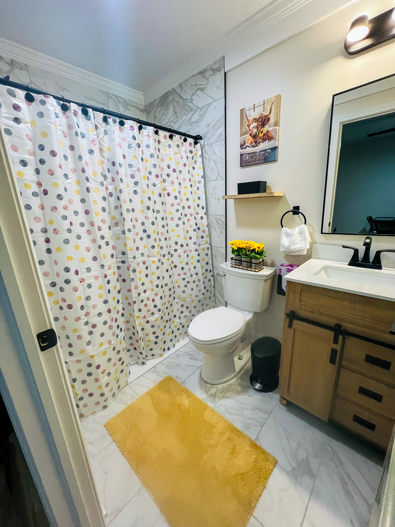 A small bathroom with a polka‑dot shower curtain, a toilet with wall shelves above it, and a vanity with a wood cabinet and black fixtures inside the home of sponsored residential provider Regina Hill in Roanoke, Virginia.