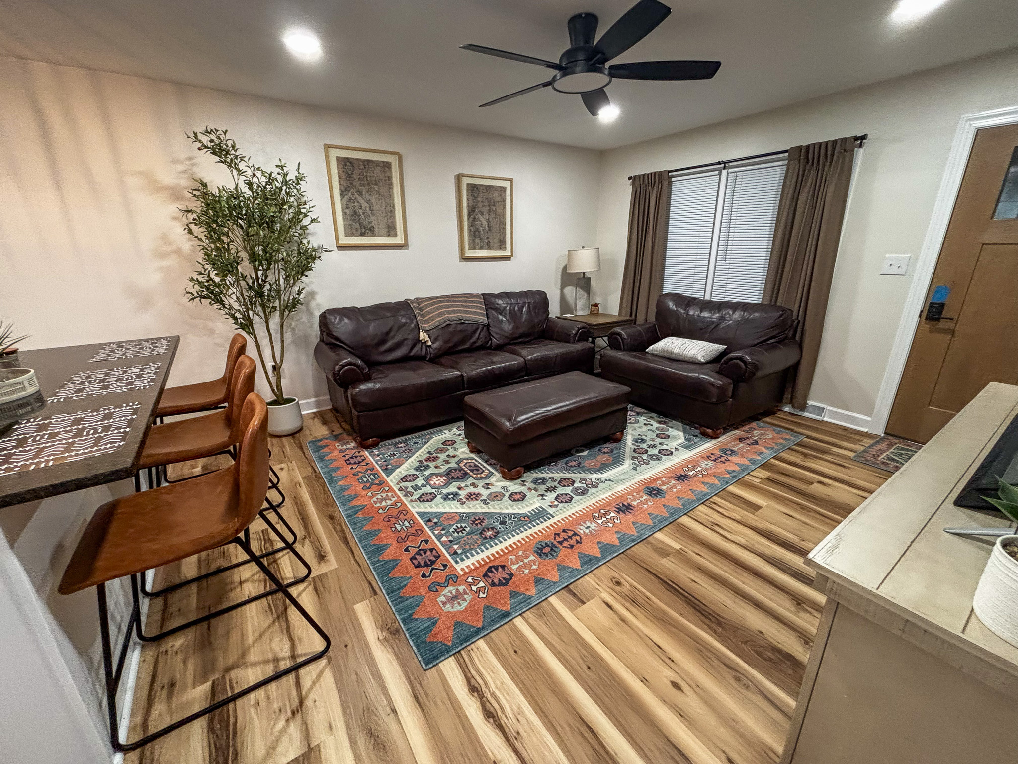 A living room with dark leather couches arranged around a patterned rug, a small ottoman, bar‑stool seating at a counter, and a large plant near the wall inside the home of sponsored residential provider Regina Hill in Roanoke, Virginia.