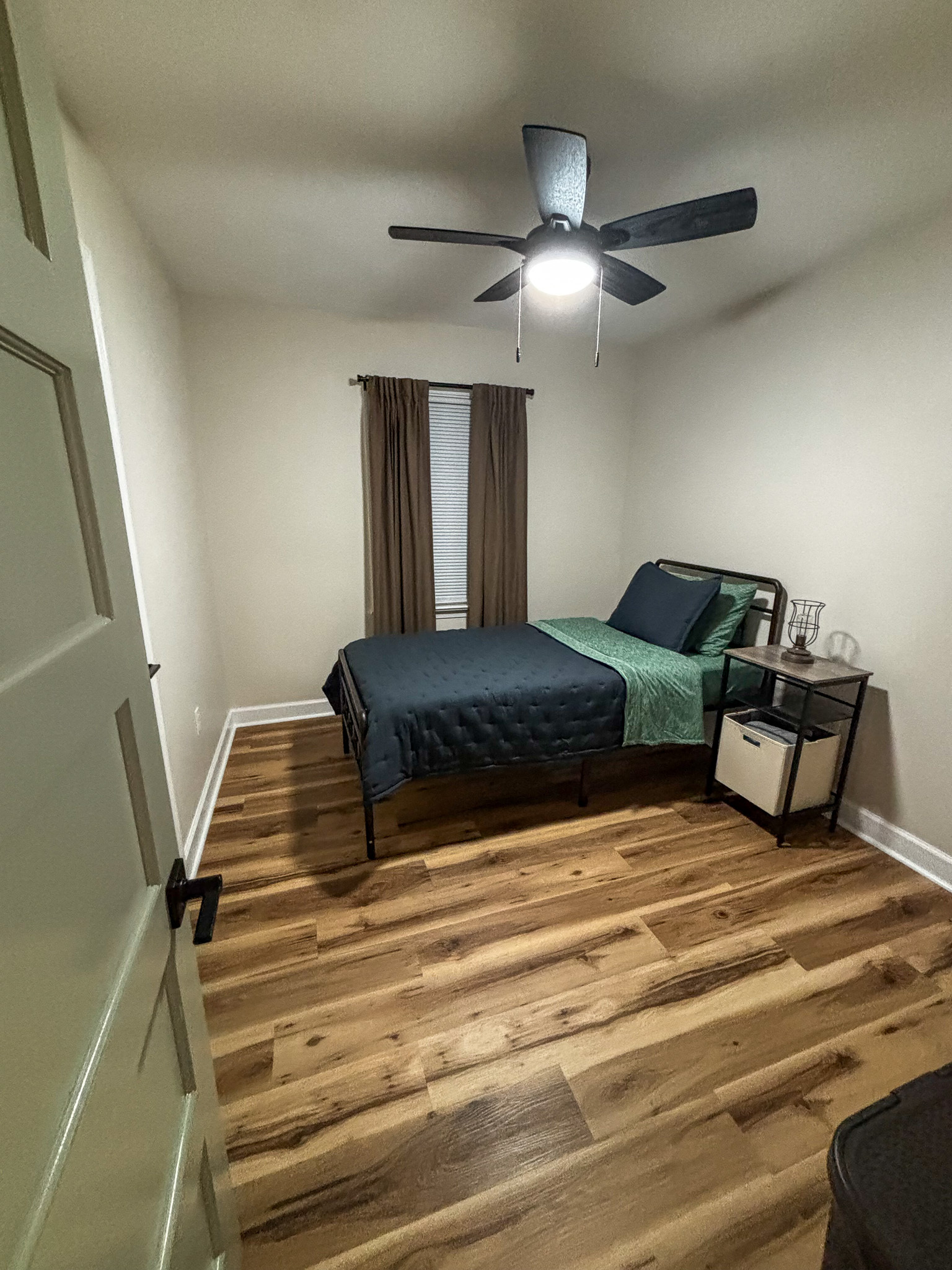 A small bedroom with wood floors, a neatly made bed with dark bedding, a nightstand holding a lamp and decor, a ceiling fan with a light, and a single window with brown curtains inside the home of sponsored residential provider Regina Hill in Roanoke, Virginia.