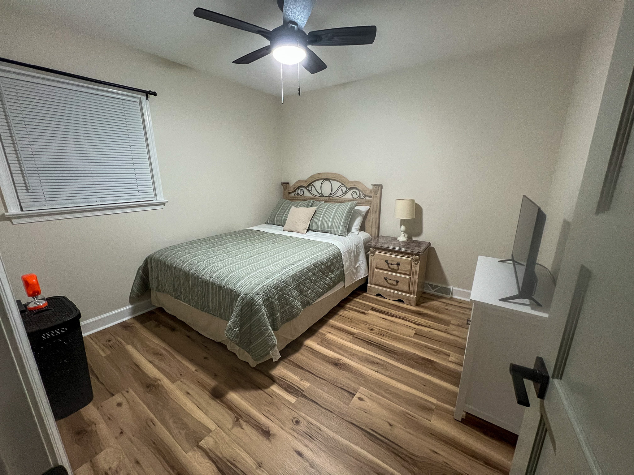 A tidy bedroom with wood floors, a neatly made bed with green bedding, a nightstand with a lamp, a ceiling fan, and a dresser with a TV inside the home of sponsored residential provider Regina Hill in Roanoke, Virginia.