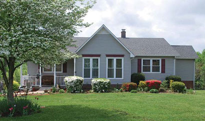 Single‑story gray house with white trim, red shutters, front porch, and well‑kept landscaping on a large green lawn belonging to Group Home Provider Rita Childress in Blairs, Virginia.