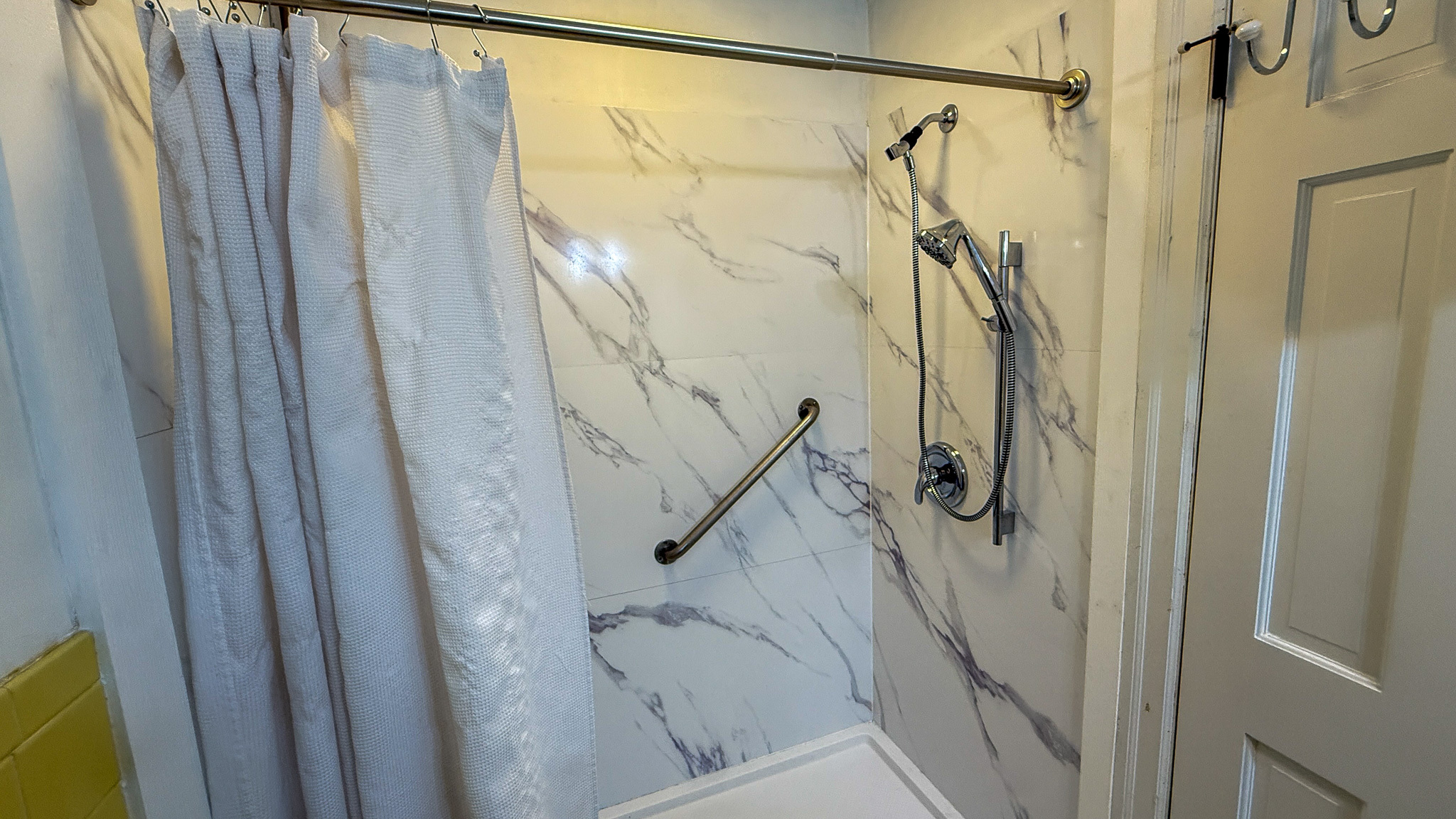 A shower area with marble‑style wall panels, a grab bar, and a handheld showerhead beside a white curtain inside the home of Sponsored Residential Providers Angela and James Snyder in Rocky Mount, Virginia.