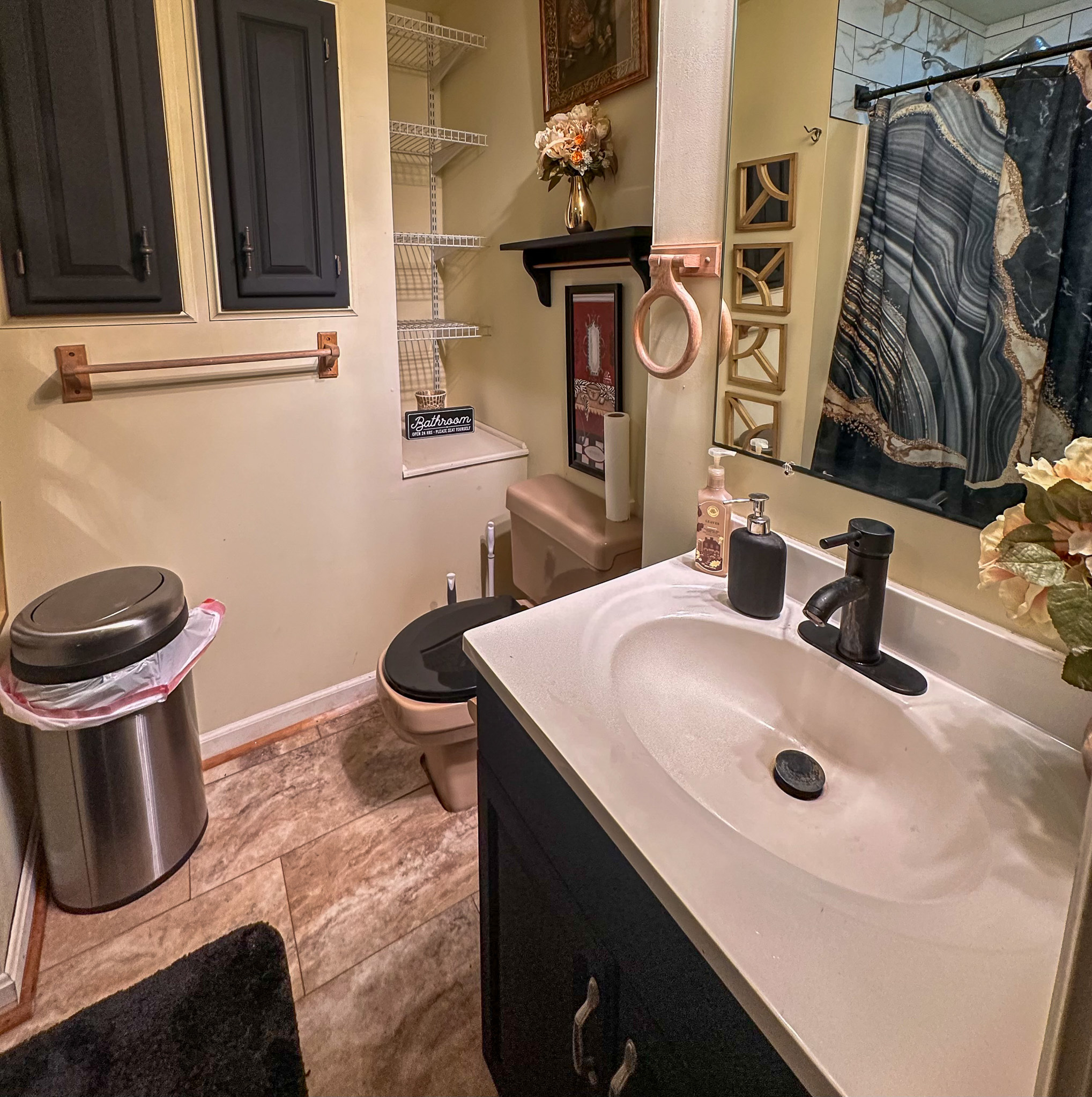 Bathroom with a white sink vanity, dark cabinets, toilet, shelving, and a dark patterned shower curtain inside the home of Sponsored Residential Provider Tarnisha Bradshaw in Fredericksburg, Virginia.