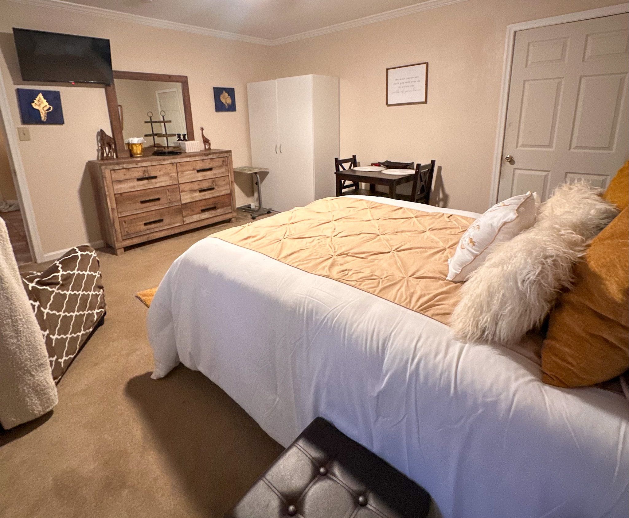 Bedroom with a bed in white and gold bedding, a wooden dresser with a wall-mounted TV, and a small dining table against the far wall inside the home of Sponsored Residential Provider Tarnisha Bradshaw.