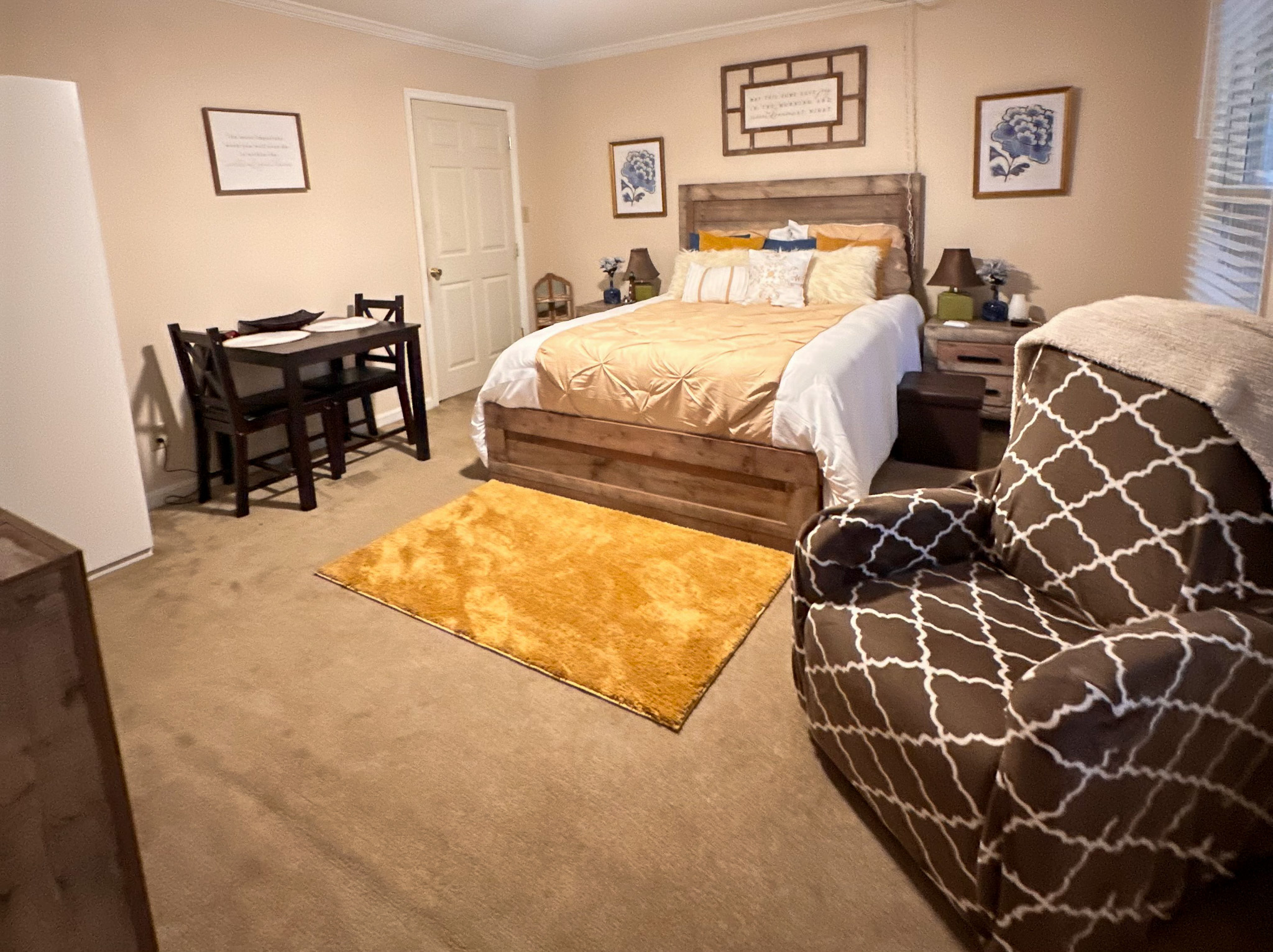 Bedroom with a wooden bed frame, gold bedding, small dining table, patterned armchair, and a yellow rug on carpeted flooring inside the home of Sponsored Residential Provider Tarnisha Bradshaw in Fredericksburg, Virginia.