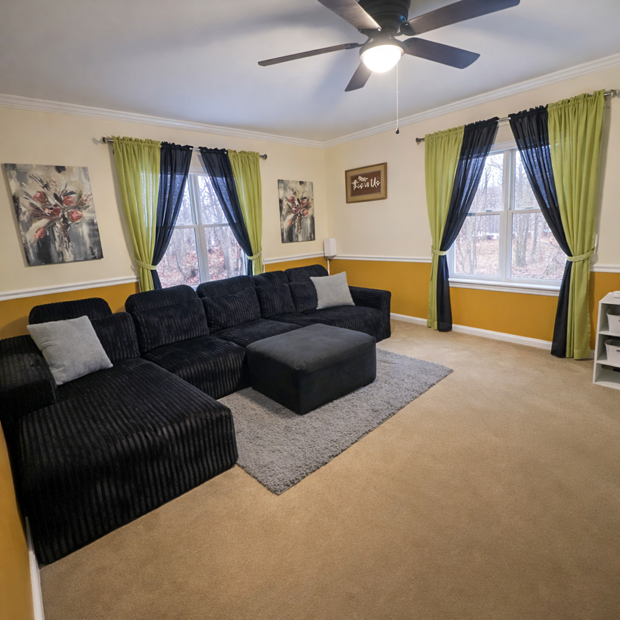 Cozy living room with a black sectional sofa, ottoman, colorful curtains, wall art, and a ceiling fan inside the home of Sponsored Residential Provider Tarnisha Bradshaw in Fredericksburg, Virginia.