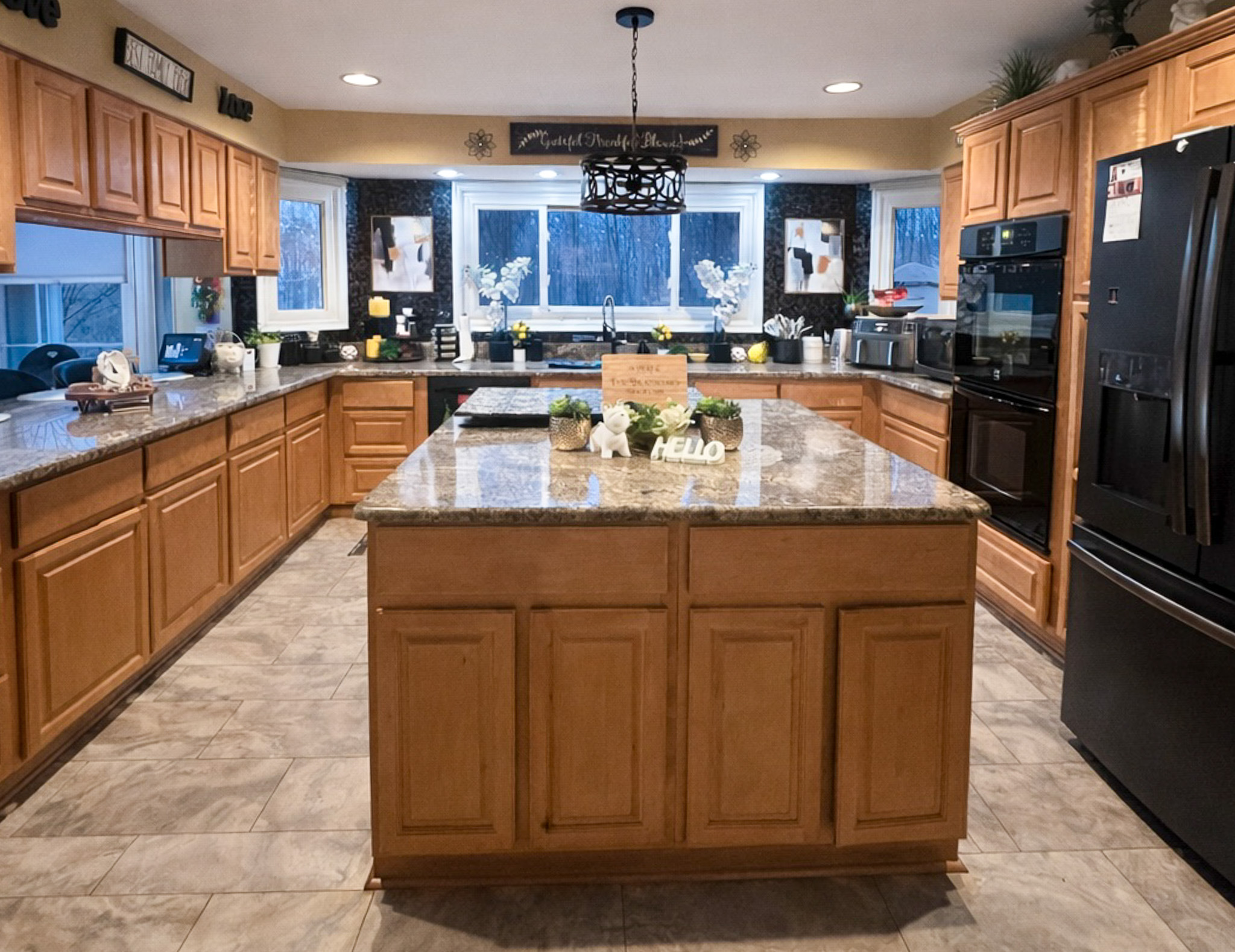 A large kitchen with light wood cabinets, a central island, black appliances, and multiple windows above the sink area inside the home of Sponsored Residential Provider Tarnisha Bradshaw in Fredericksburg, Virginia.