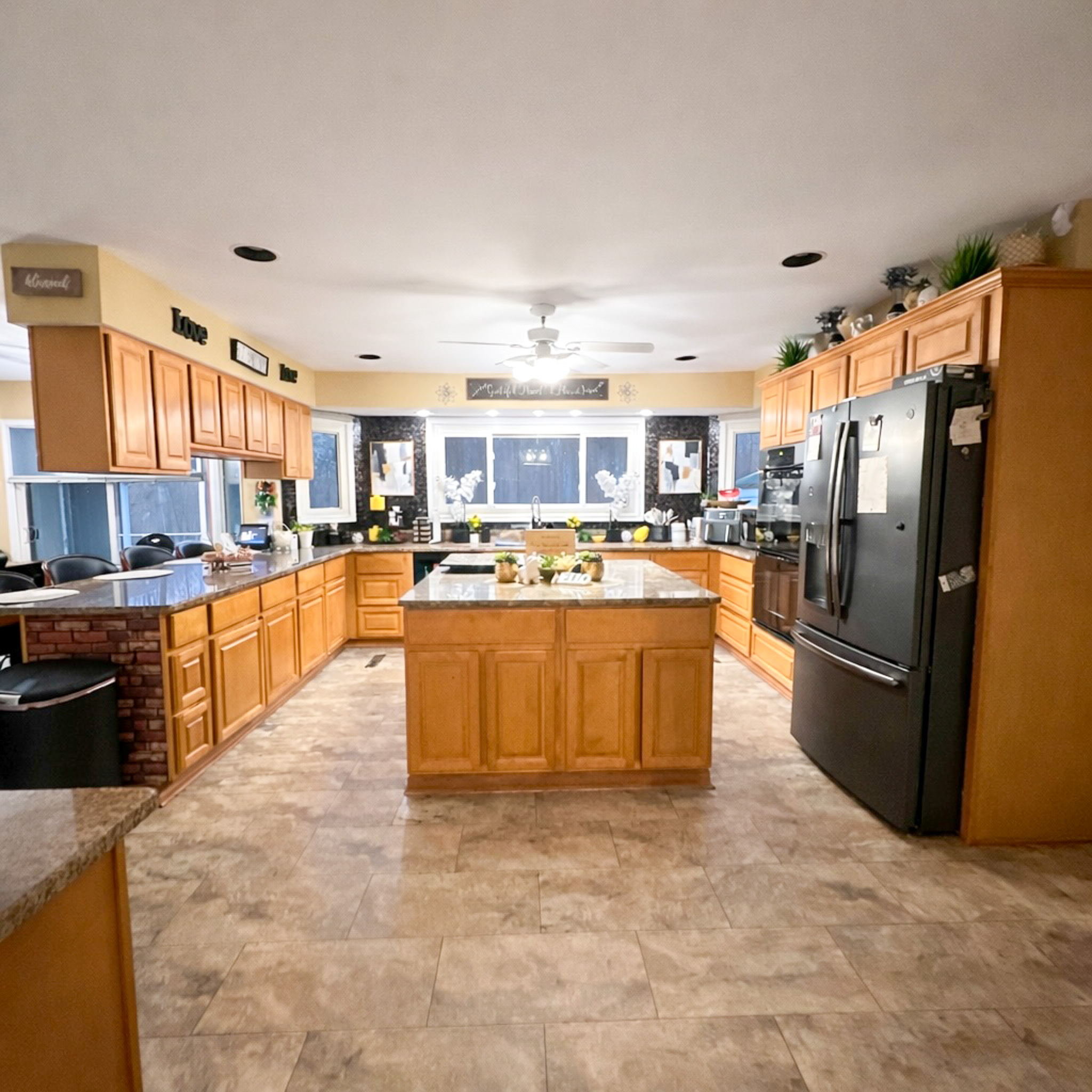 A spacious kitchen with light wood cabinets, a central island, black appliances, and a row of windows above the sink area inside the home of Sponsored Residential Provider Tarnisha Bradshaw in Fredericksburg, Virginia.
