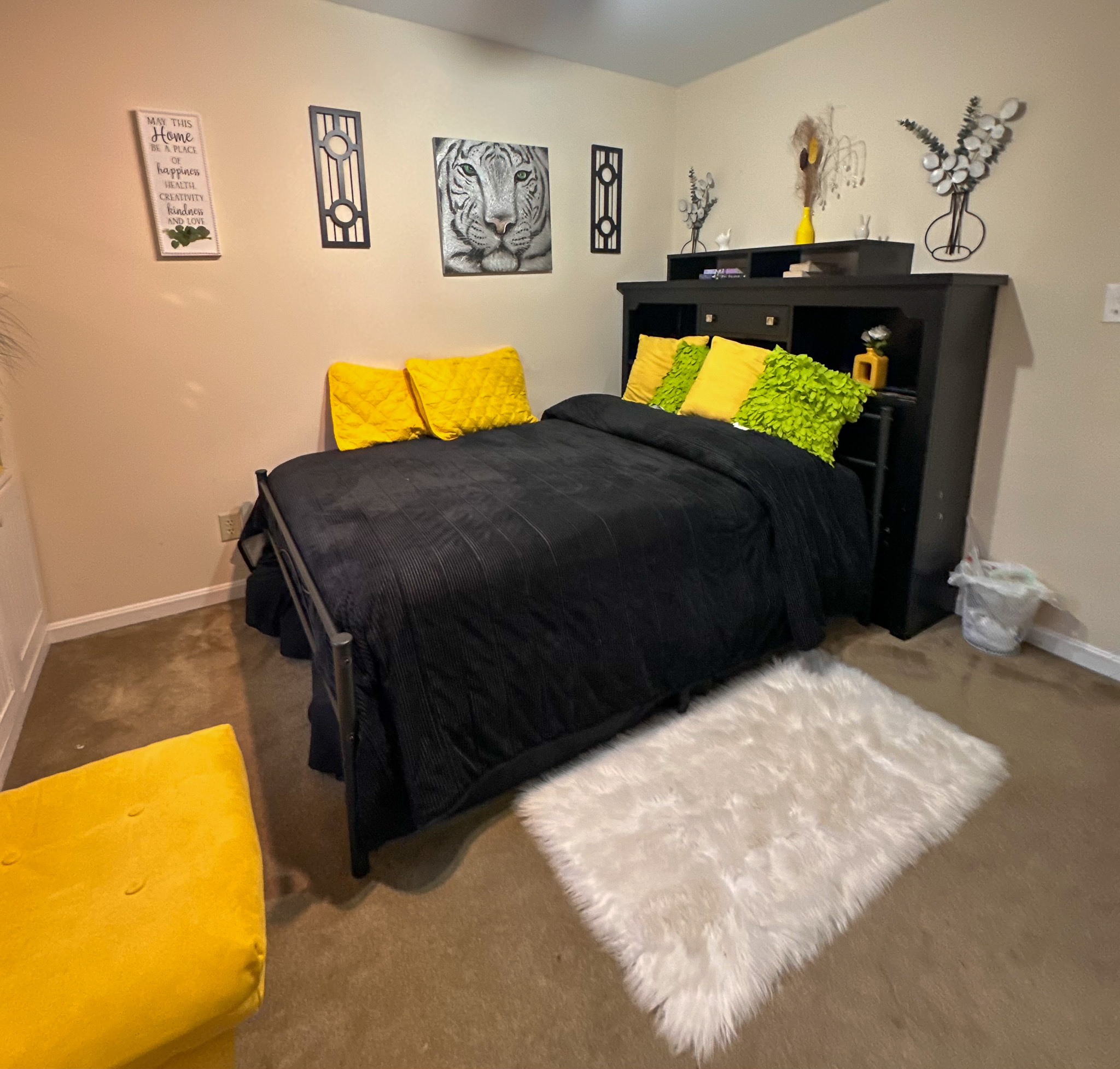 A bedroom with a bed dressed in black bedding and bright yellow and green accent pillows, a white fluffy rug, a black headboard with shelves, and wall art above the bed inside the home of Sponsored Residential Provider Tarnisha Bradshaw in Fredericksburg, Virginia.