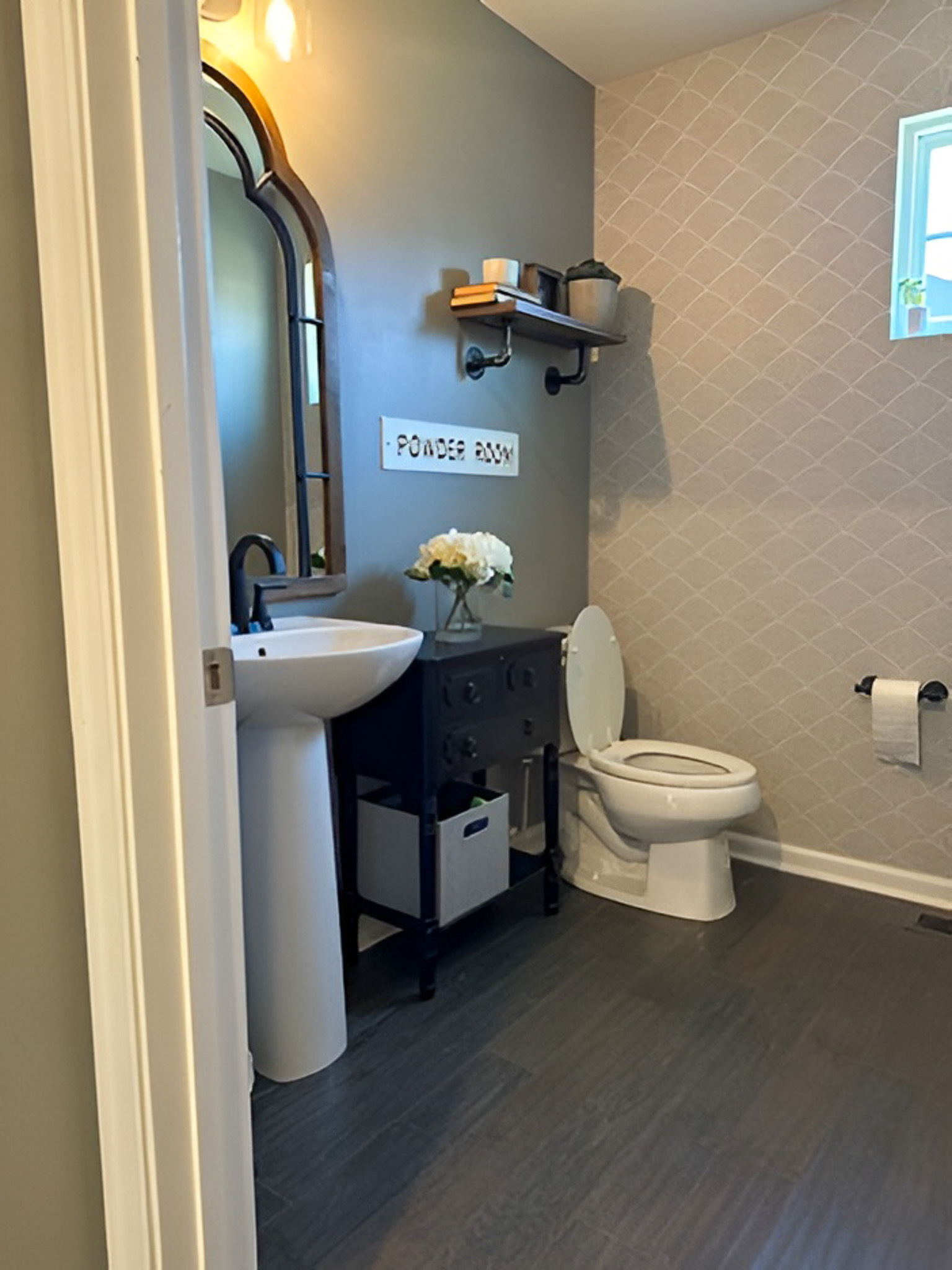 Small bathroom with a pedestal sink, toilet, wall shelf, and decorative mirror inside the home of Group Home Providers Alex and Dillon Tittle in Fredericksburg, Virginia.