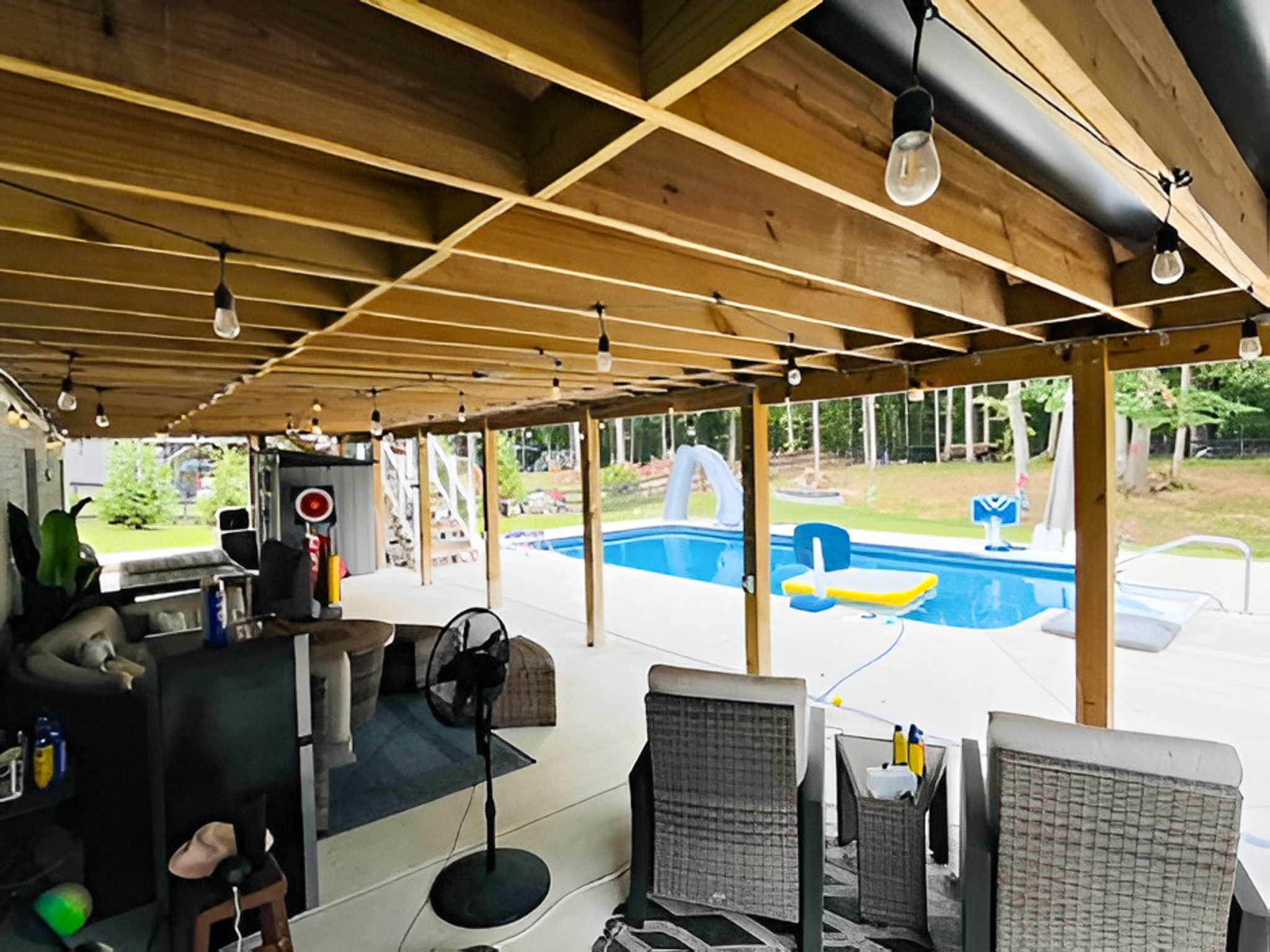 Covered patio with exposed wooden beams, string lights, seating area, and a view of a backyard pool at the home of Group Home Providers Alex and Dillon Tittle in Fredericksburg, Virginia.