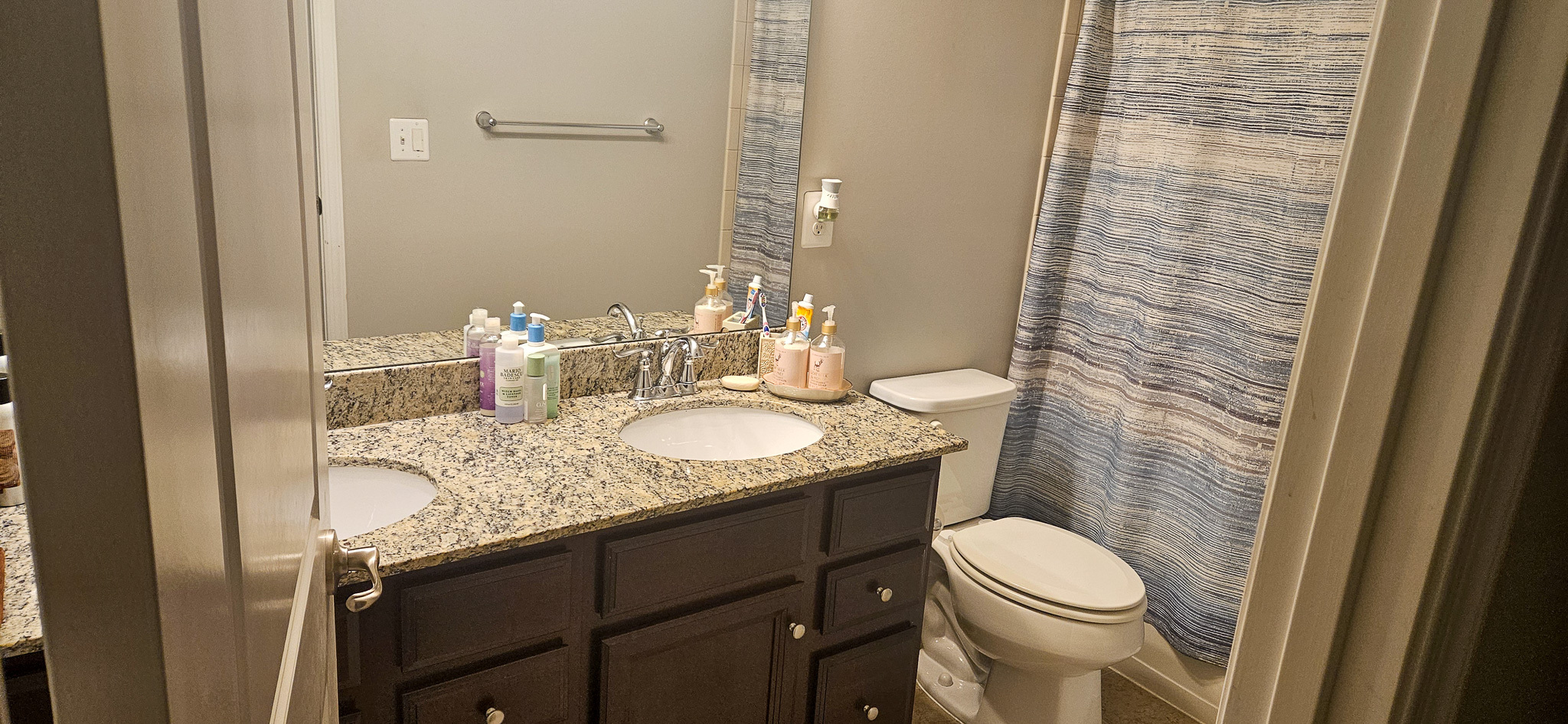 A bathroom with a granite double-sink vanity, assorted toiletries on the counter, a toilet, and a blue-striped shower curtain inside the home of Sponsored Residential Provider Vanessa Moore in Fredericksburg, Virginia.