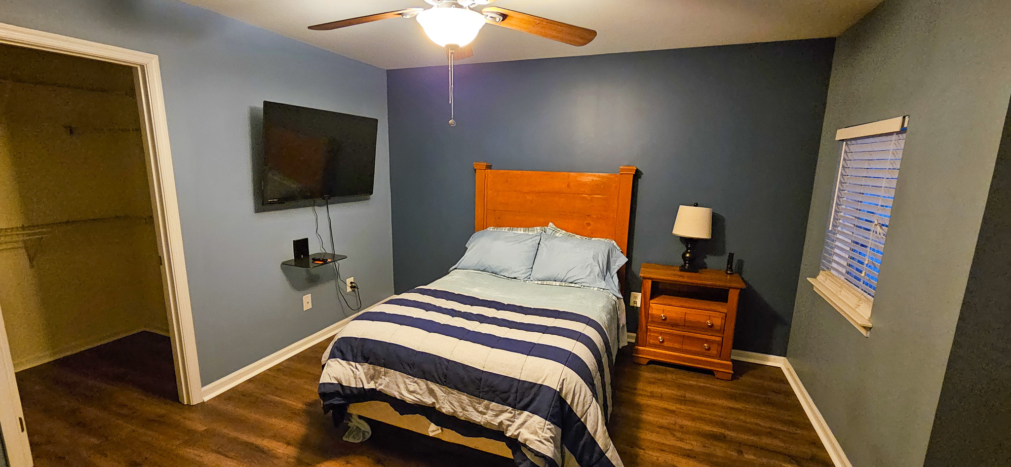 A bedroom with a wooden bed frame, striped bedding, a nightstand with a lamp, a wall‑mounted TV, and an open walk‑in closet inside the home of Sponsored Residential Provider Vanessa Moore in Fredericksburg, Virginia.