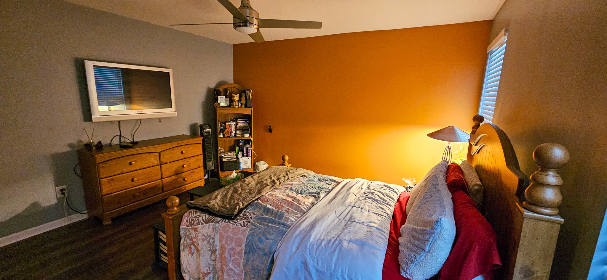 A bedroom with an orange accent wall, a wooden dresser with a TV above it, a bookshelf, and a bed with layered bedding and pillows inside the home of Sponsored Residential Provider Vanessa Moore in Fredericksburg, Virginia.