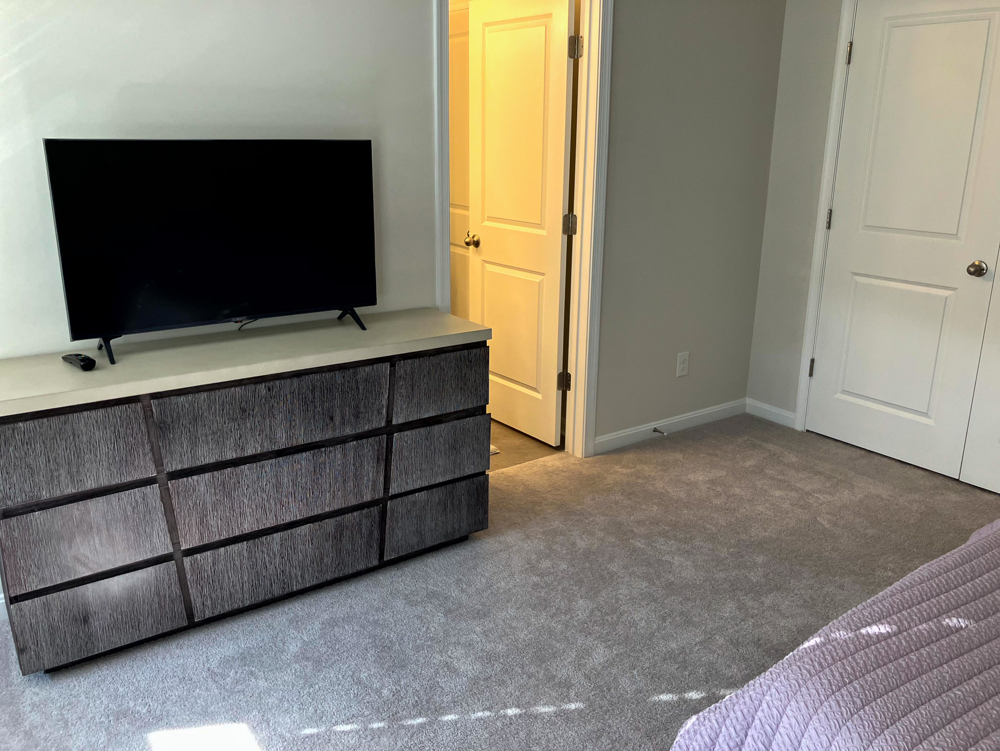 Bedroom with a dark wood dresser holding a TV, an open doorway to a lit bathroom, and two closed white doors inside the home of Sponsored Residential Provider Homaira Yazdani in Fredericksburg, Virginia.