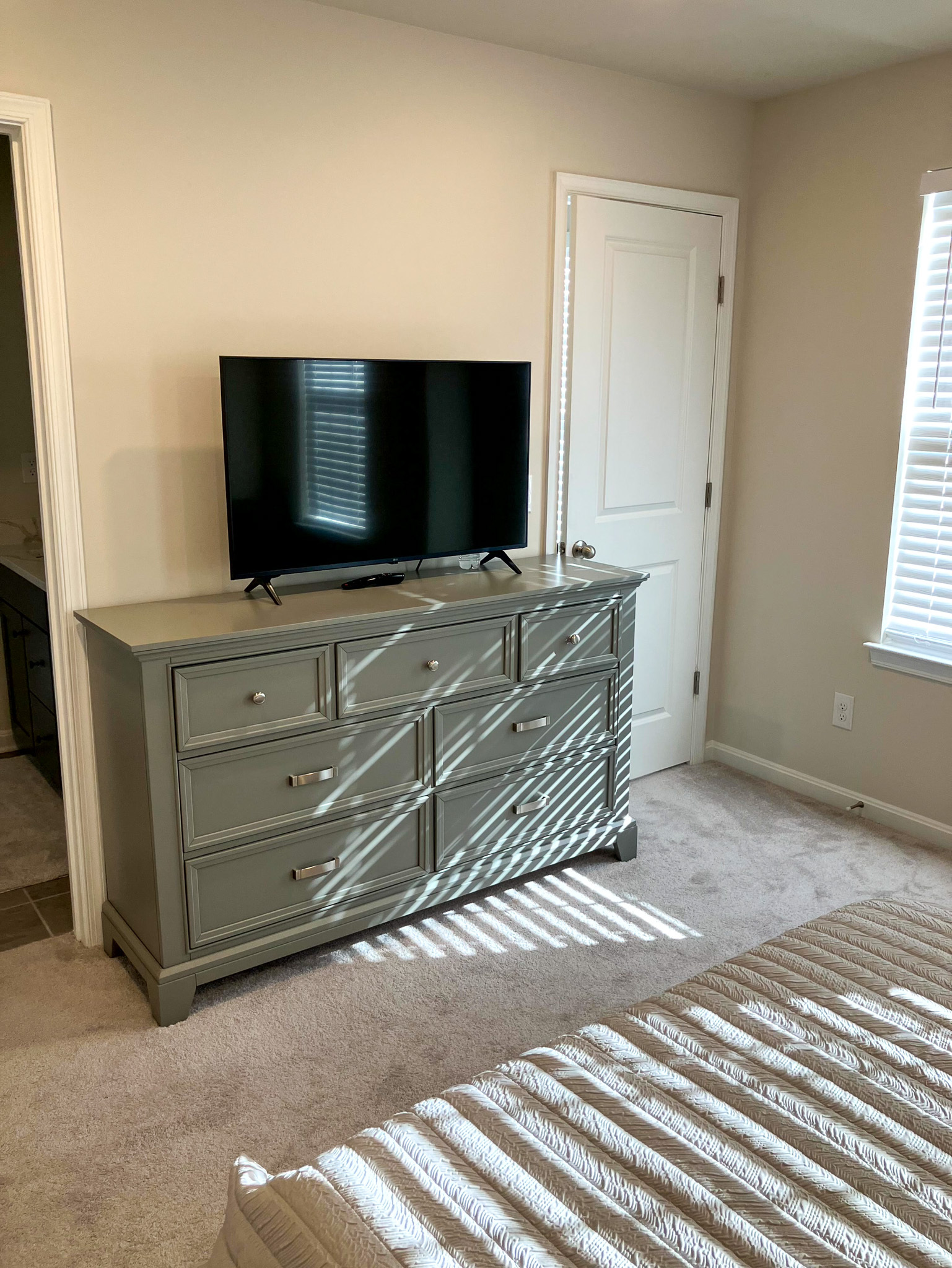 Bedroom dresser with a TV on top, a doorway to a bathroom, a closed white door, and sunlight casting shadows across the carpet inside the home of Sponsored Residential Provider Homaira Yazdani in Fredericksburg, Virginia.