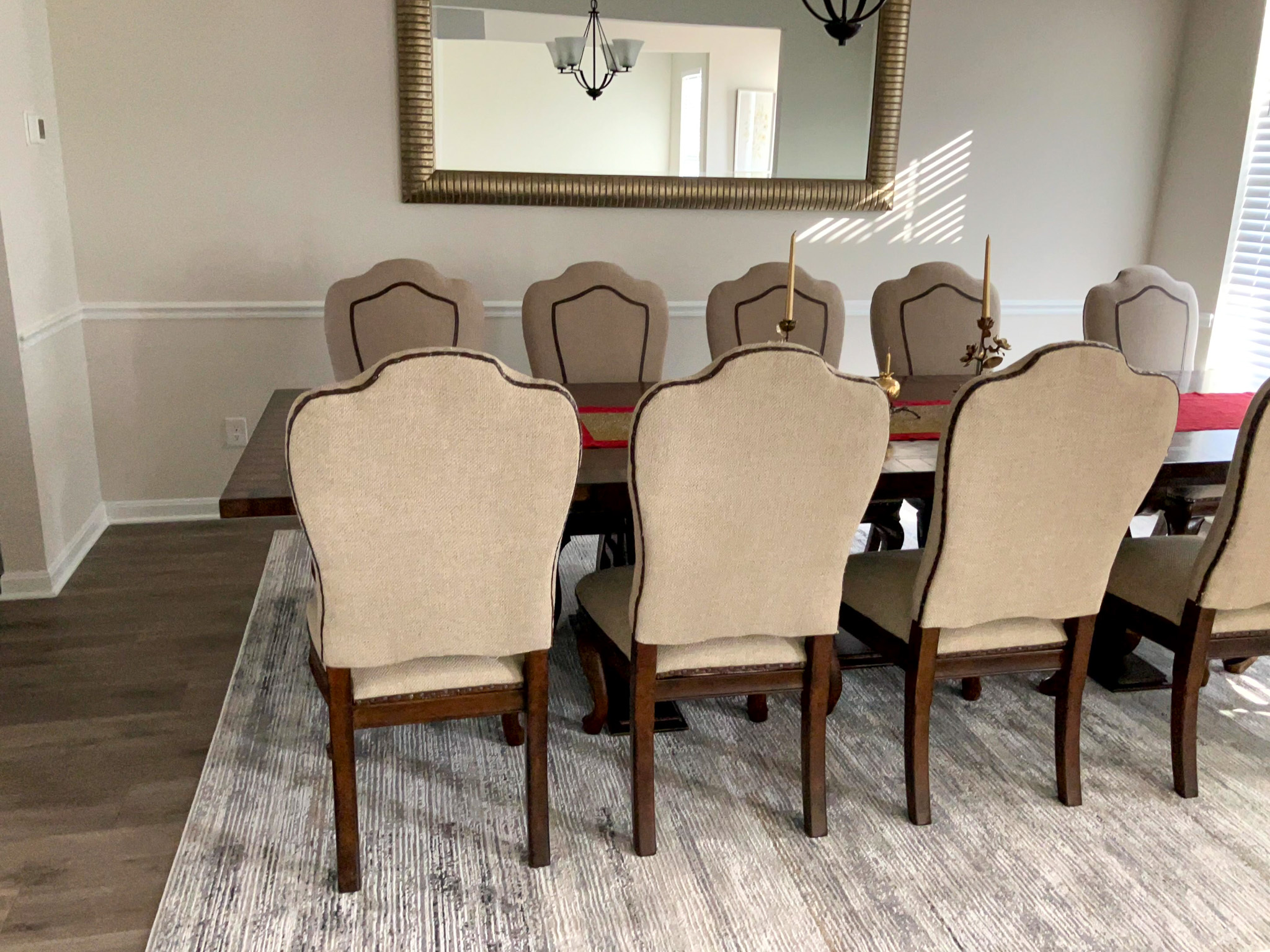 Dining room with a long wooden table, beige upholstered chairs, a large wall mirror, and a gray area rug  inside the home of Sponsored Residential Provider Homaira Yazdani in Fredericksburg, Virginia.