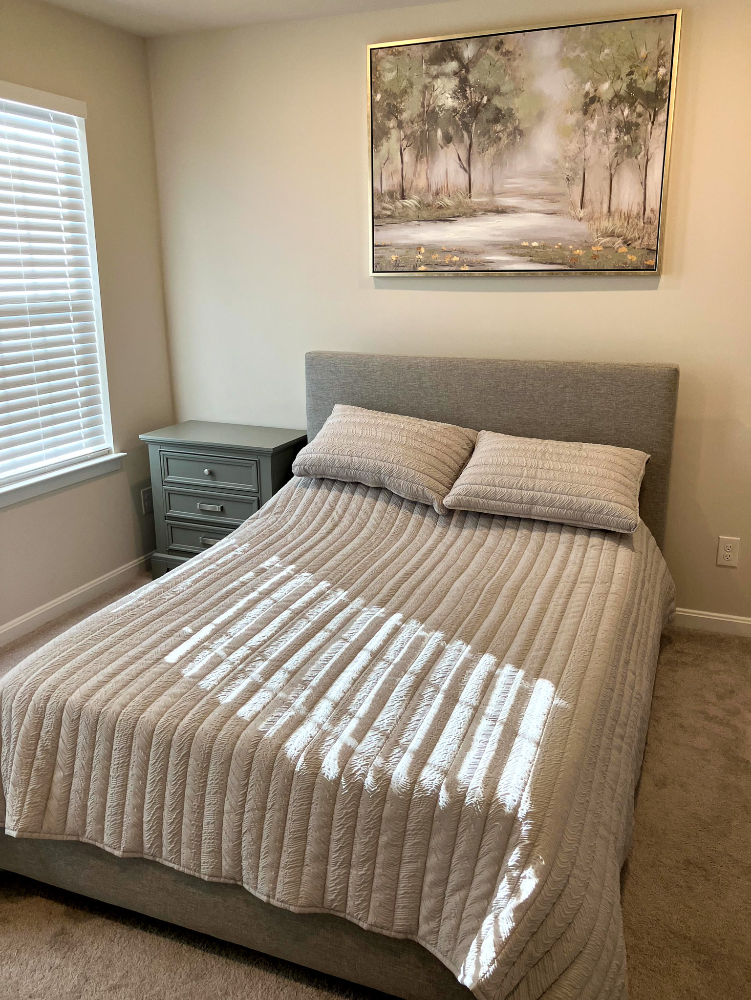 Bedroom with a neatly made bed in neutral bedding, a nightstand to the left, and a landscape painting above the headboard inside the home of Sponsored Residential Provider Homaira Yazdani in Fredericksburg, Virginia.