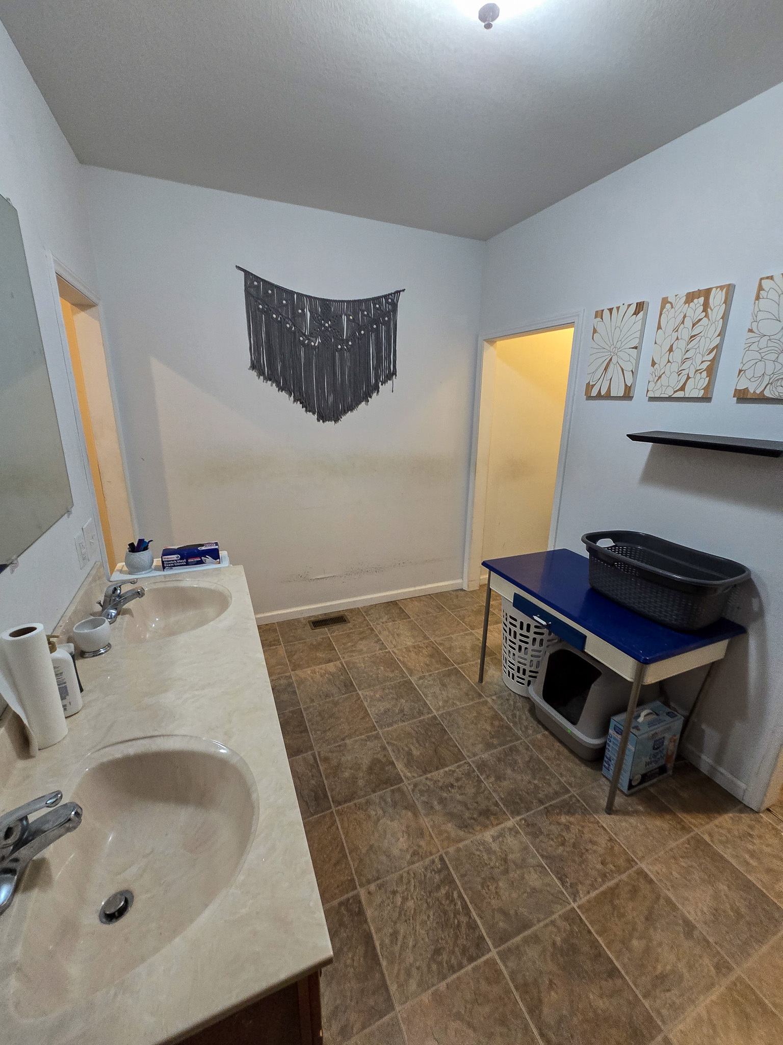 A simple bathroom with a double sink vanity, tile flooring, light walls, wall art, and a small table holding laundry items near two open doorways inside the home of Sponsored Residential Provider Chris Sumner in Galax, Virginia.