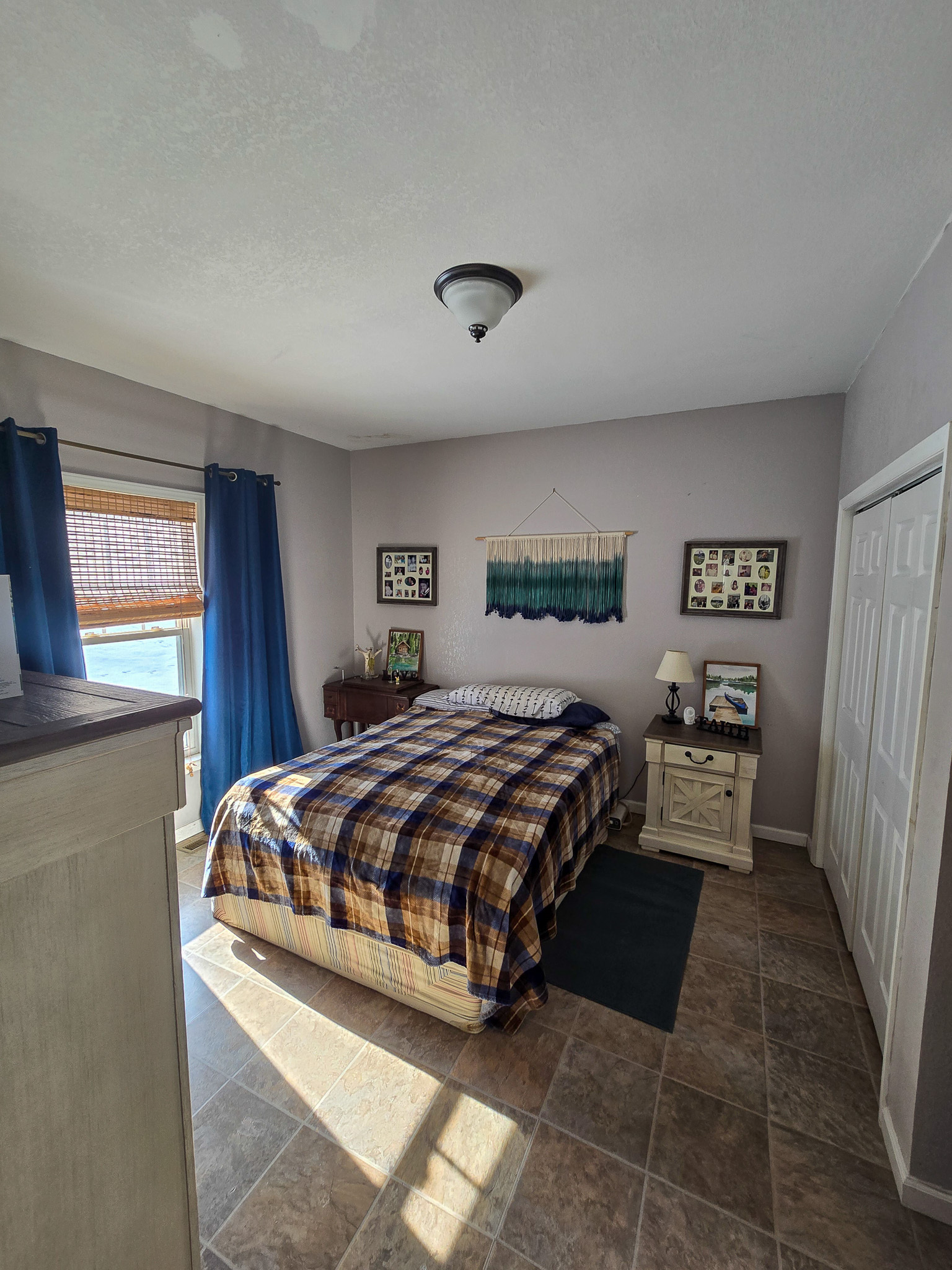 A bright bedroom with a plaid‑patterned bed, blue curtains, light‑colored walls, tile flooring, and a nightstand with décor beneath wall art inside the home of Sponsored Residential Provider Chris Sumner in Galax, Virginia.