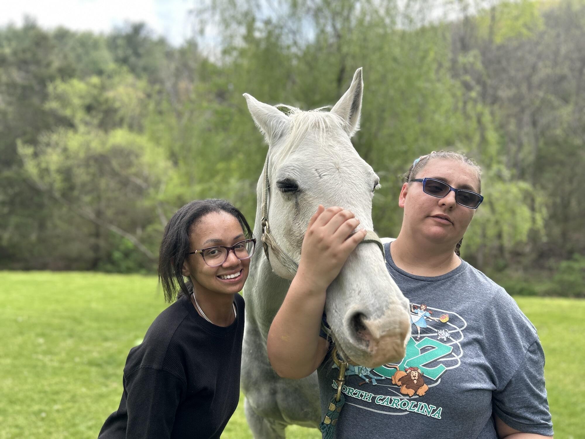 A girl standing outside in the grass with her arm around a white horse alongside a therapeutic recreation consultant