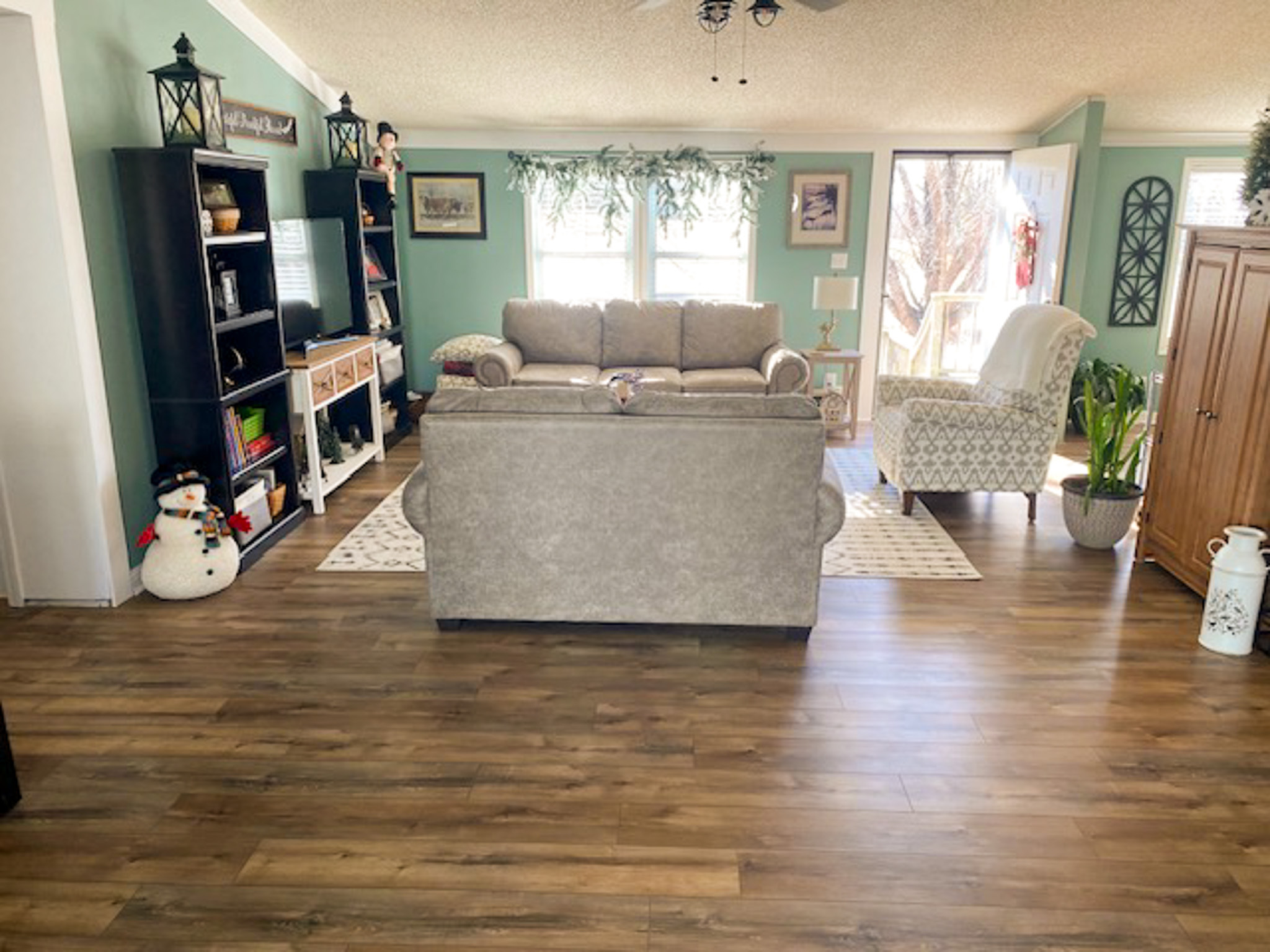 A bright living room with mint‑green walls, wood‑look floors, a sofa facing a TV on dark shelving, and additional seating near large windows and a sliding glass door inside the home of Sponsored Residential Provider Christina Bedsaul in Hillsville, Virginia.