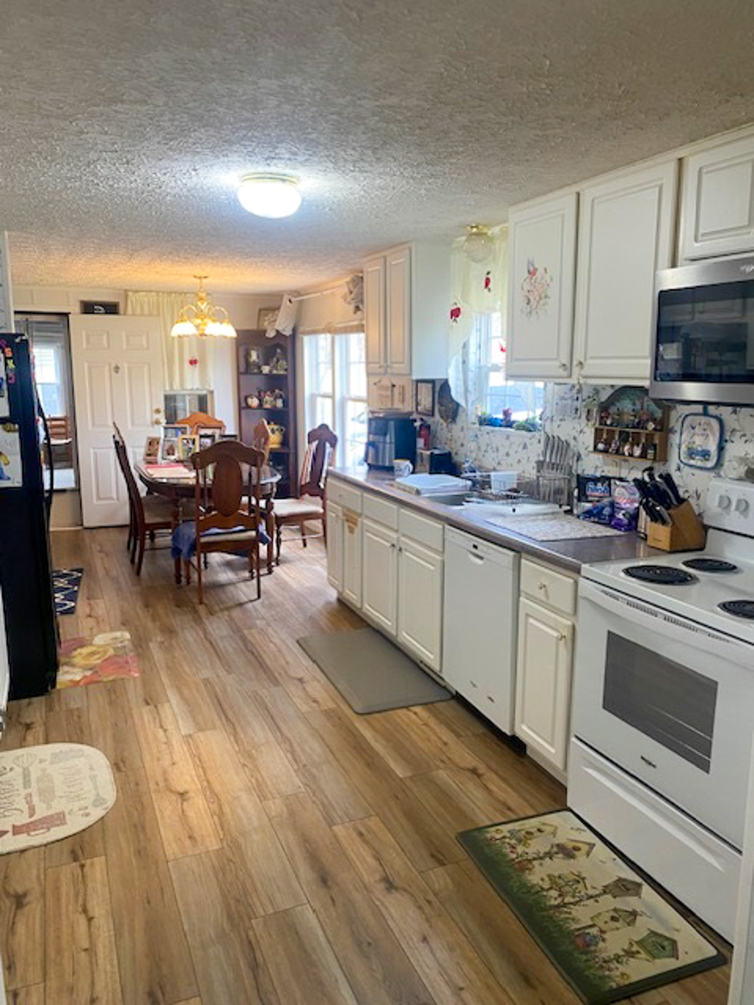 Kitchen with white cabinets and appliances, wood‑look flooring, and a dining area with a wooden table in the background inside the home of Sponsored Residential Provider Angel Dixon in Woodlawn, Virginia.