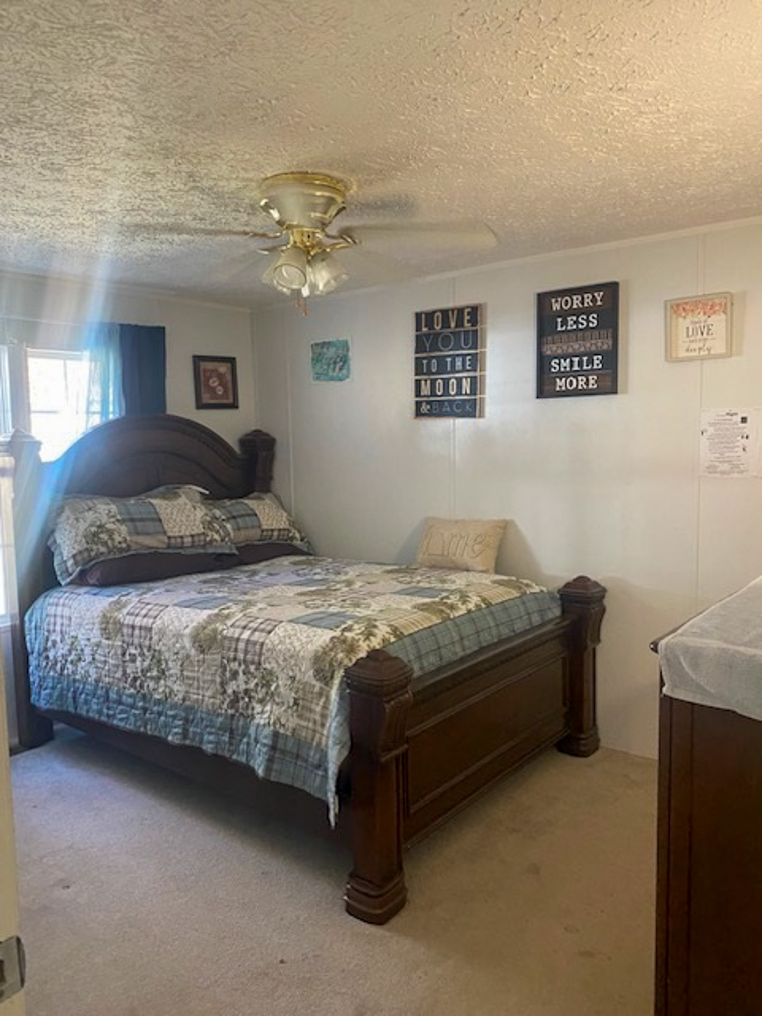 Bedroom with a wooden bed frame, patterned bedding, wall art, a ceiling fan, and a small window with dark curtains inside the home of Sponsored Residential Provider Angel Dixon in Woodlawn, Virginia.