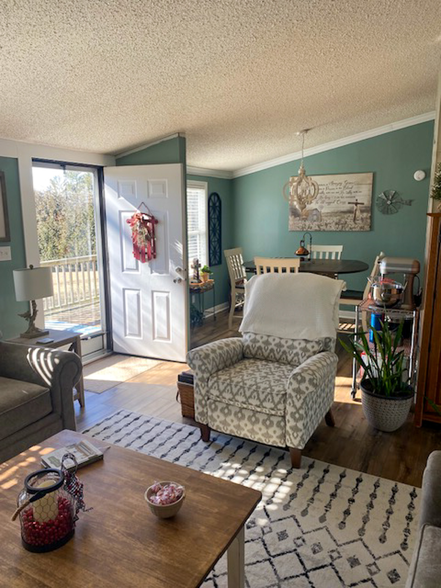 A cozy living space with teal walls, patterned armchairs, a wooden coffee table, and an open front door letting in sunlight, with a small dining area visible in the background inside the home of Sponsored Residential Provider Christina Bedsaul in Hillsville, Virginia.