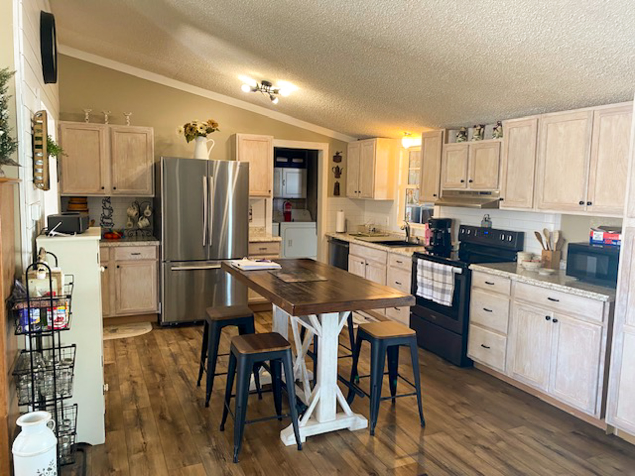 A bright kitchen with light wood cabinets, stainless‑steel appliances, wood‑look flooring, and a central island with a wooden top and four stools inside the home of Sponsored Residential Provider Christina Bedsaul in Hillsville, Virginia.
