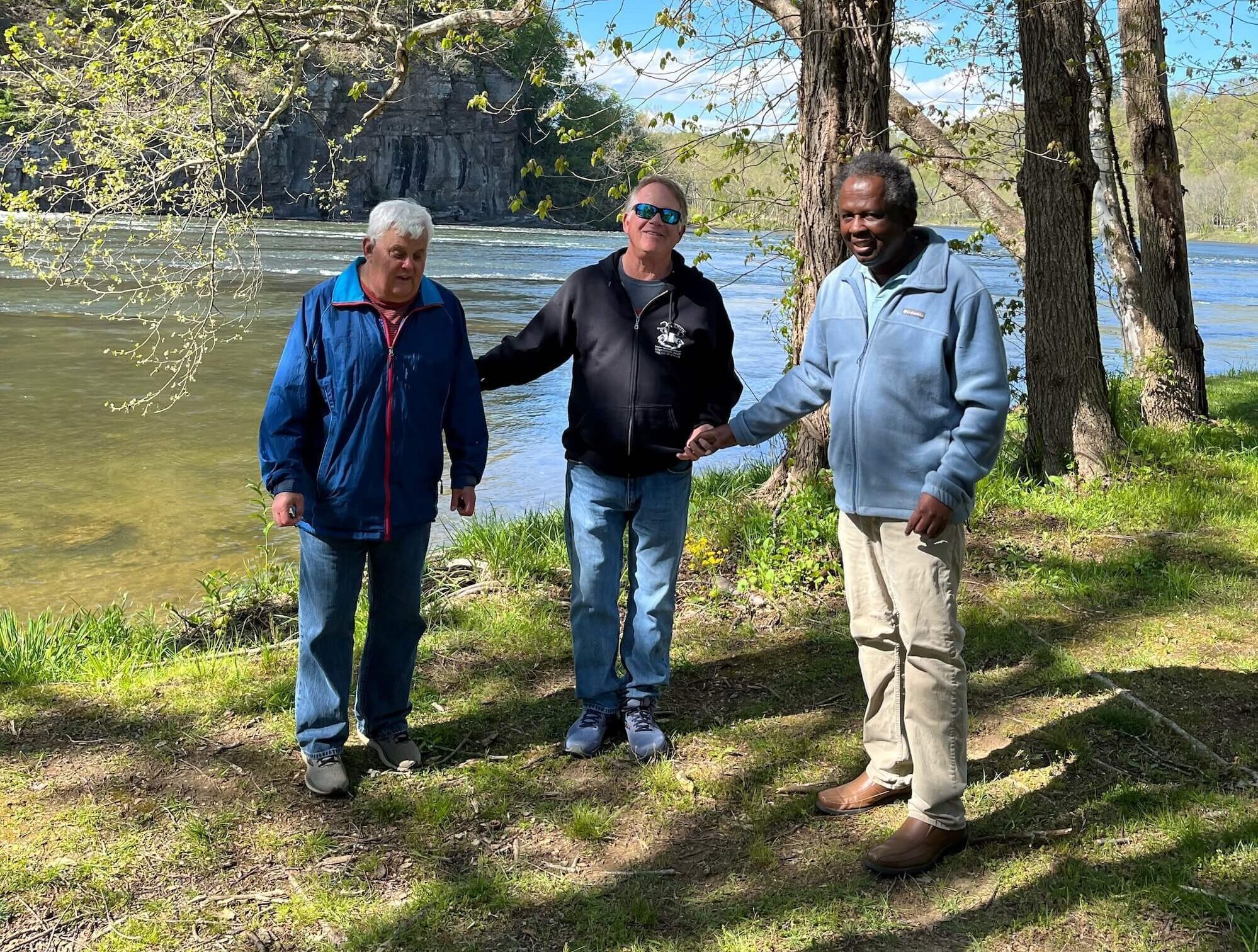 Three men standing together in front of a lake and trees