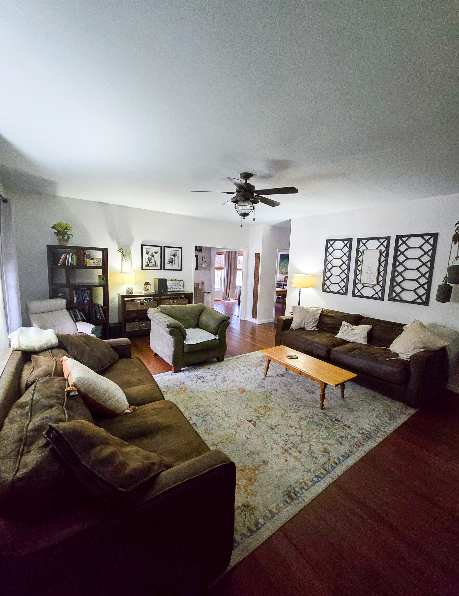 A cozy living room with two large brown sofas, an armchair, a patterned area rug, and wall art. A ceiling fan hangs above, and warm lamps illuminate bookshelves and side tables inside the home of Sponsored Residential Provider Chris Sumner in Galax, Virginia.