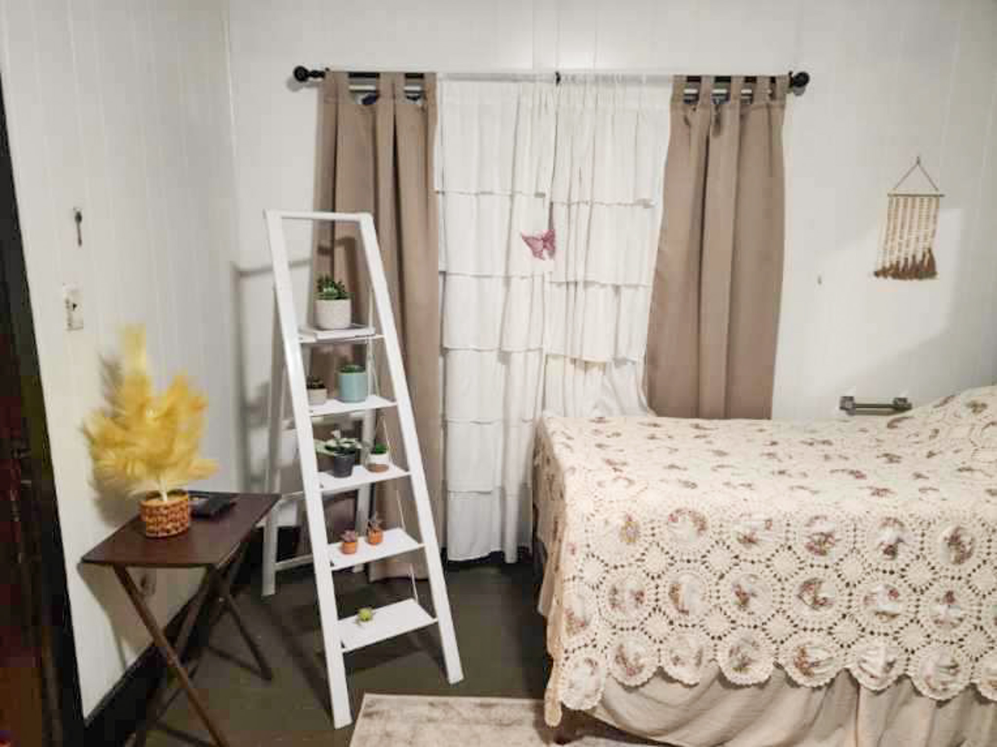 Bedroom corner with a bed covered in a lace bedspread, beige curtains, a white ladder shelf with plants, and a small side table with decor inside the home of Sponsored Residential Provider Angela Justice in Mathews, Virginia.