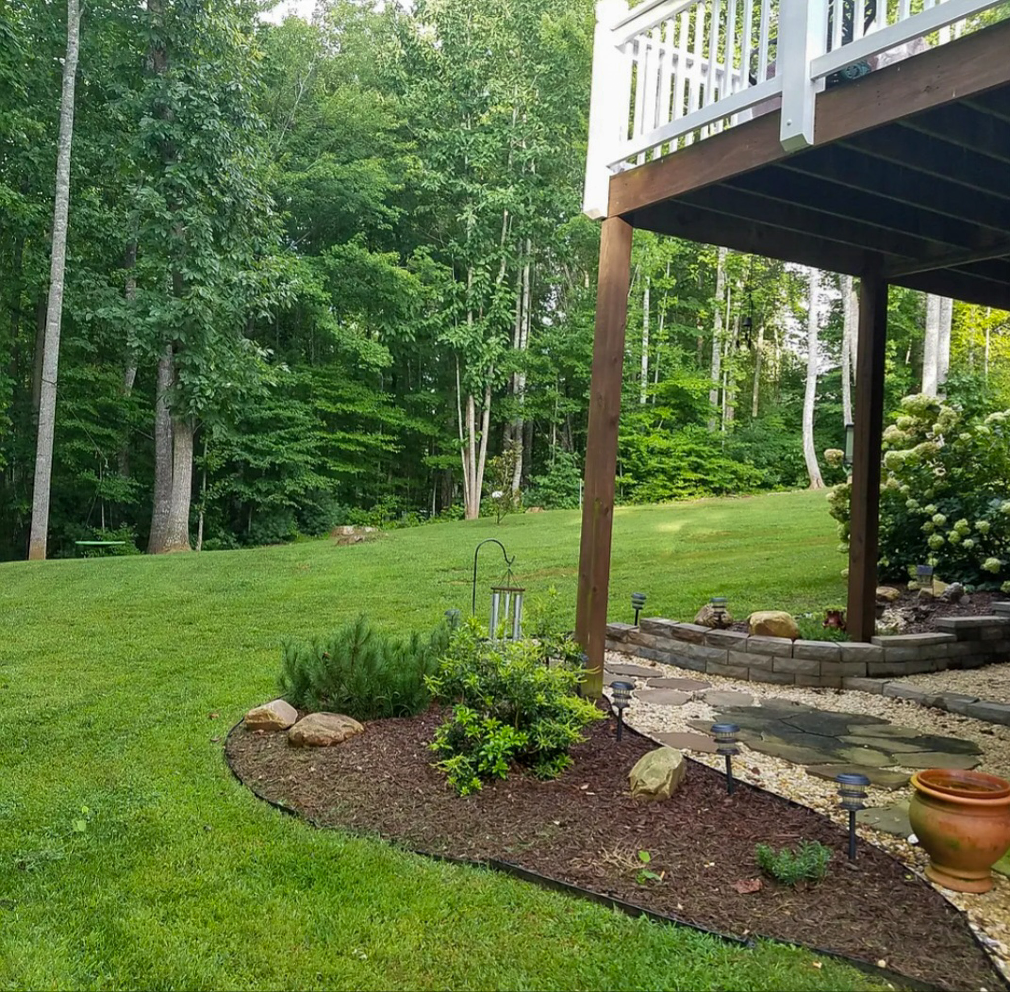 A landscaped backyard with green grass, a mulched garden bed, stone path, and the underside of a raised deck overlooking a wooded area at the home of Sponsored Residential Provider Chris Sumner in Galax, Virginia.