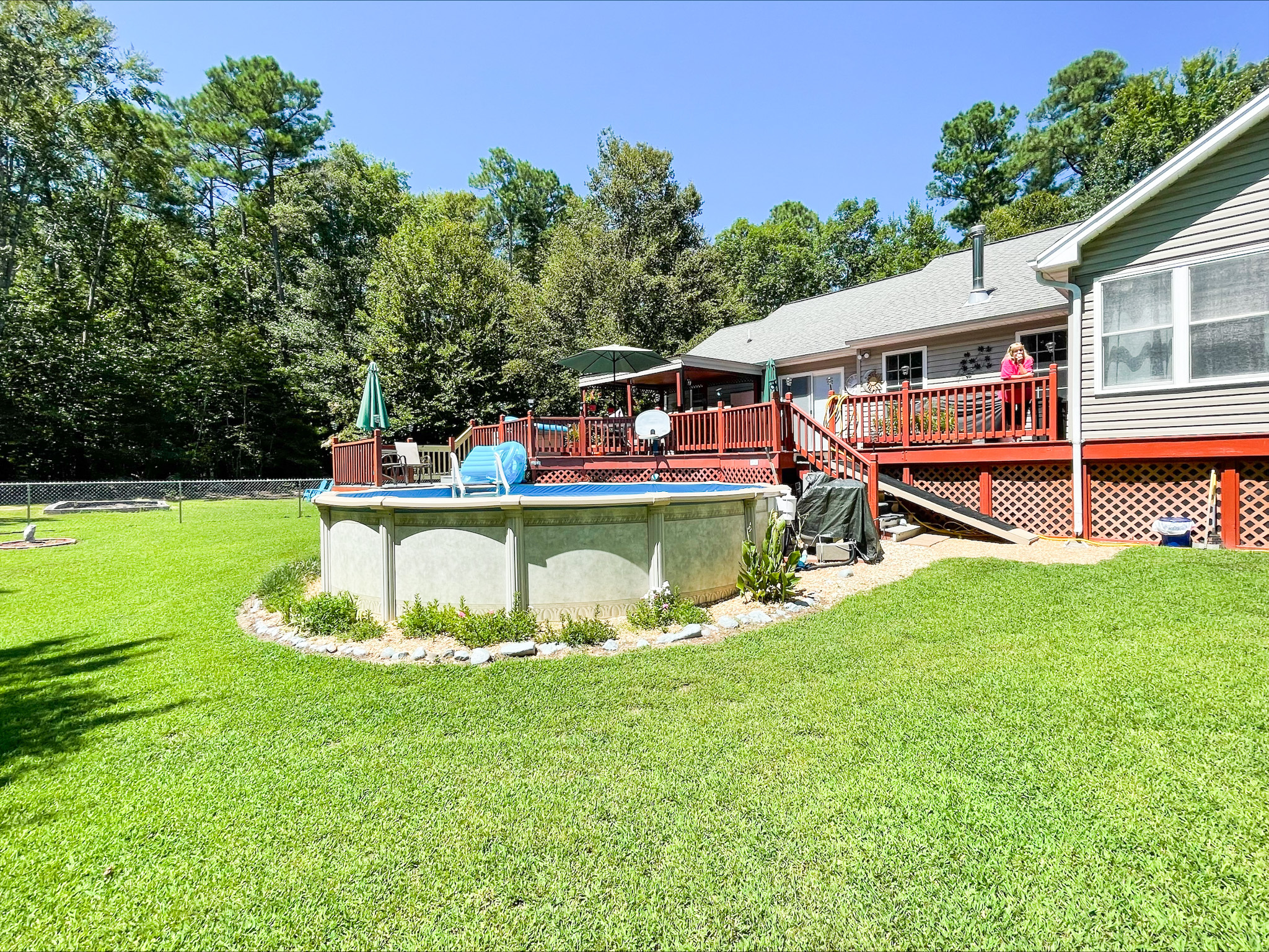 Backyard view of a house with a large wooden deck and an above‑ground pool, surrounded by a wide grassy lawn and trees outside the home of Sponsored Residential Provider Kellie Lynch in Gloucester, Virginia.