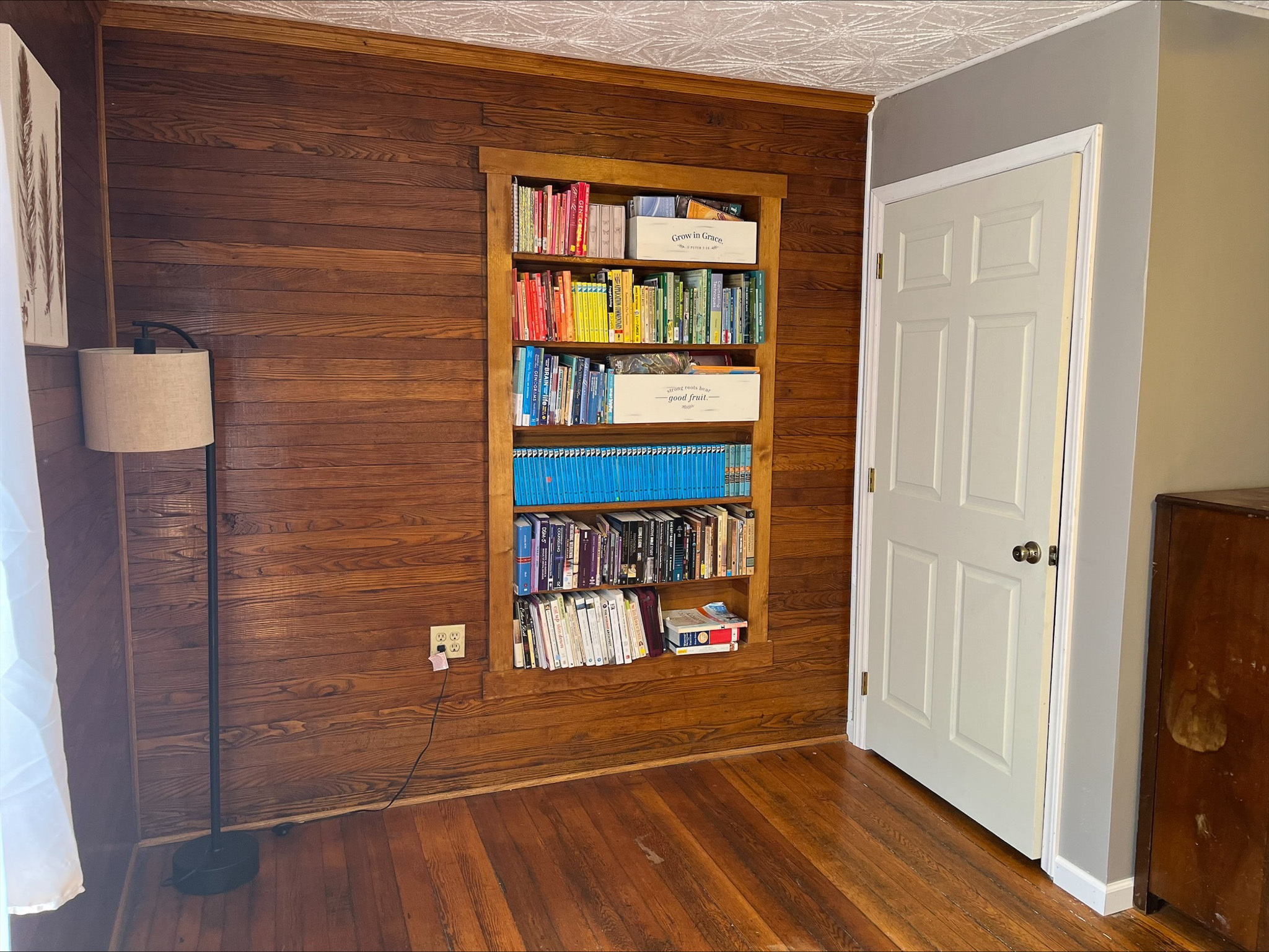 Built‑in bookshelf filled with colorful books set into a wood-paneled wall beside a white door and a floor lamp inside the home of Sponsored Residential Provider Dara Short in Willis, Virginia.