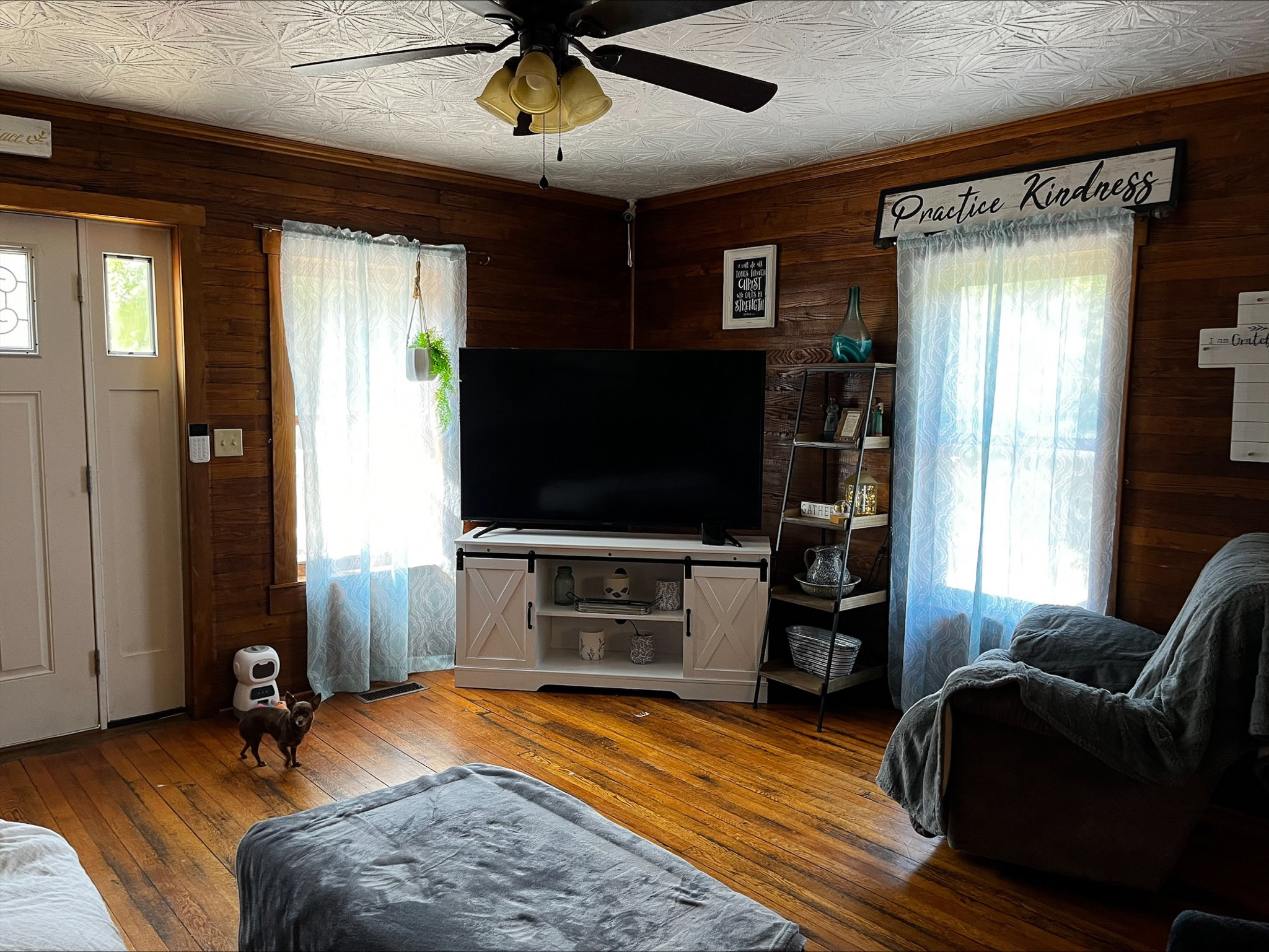Cozy living room with wood-paneled walls, a large TV on a white stand, armchair, ottoman, and two windows with sheer curtains inside the home of Sponsored Residential Provider Dara Short in Willis, Virginia.
