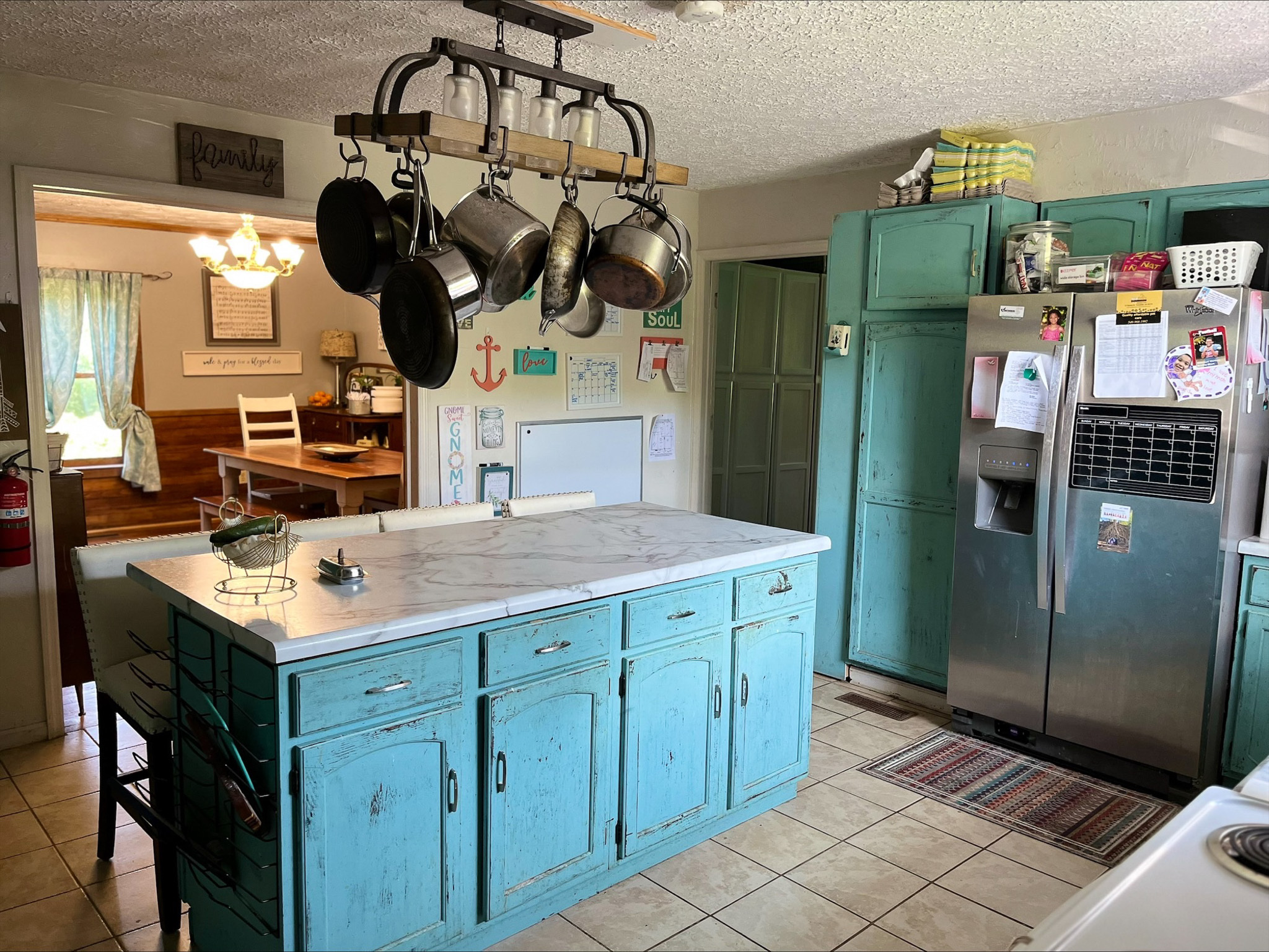 Kitchen with turquoise cabinets and island, stainless-steel fridge, tile floor, and hanging pot rack, with a view into the dining room inside the home of Sponsored Residential Provider Dara Short in Willis, Virginia.