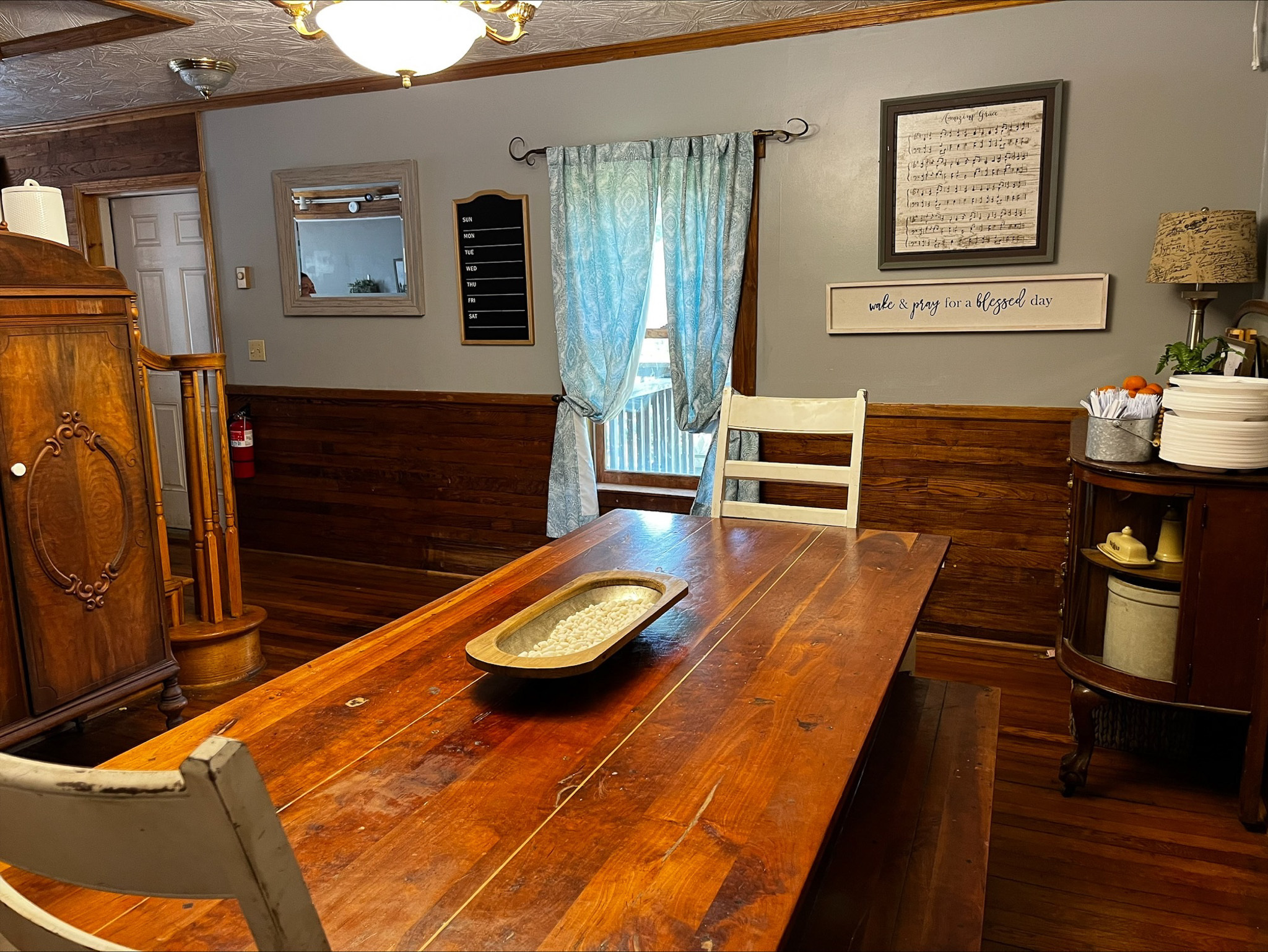 Dining room with a wooden table, two chairs, blue curtains over a window, and wall décor above wood-paneled wainscoting inside the home of Sponsored Residential Provider Dara Short in Willis, Virginia.