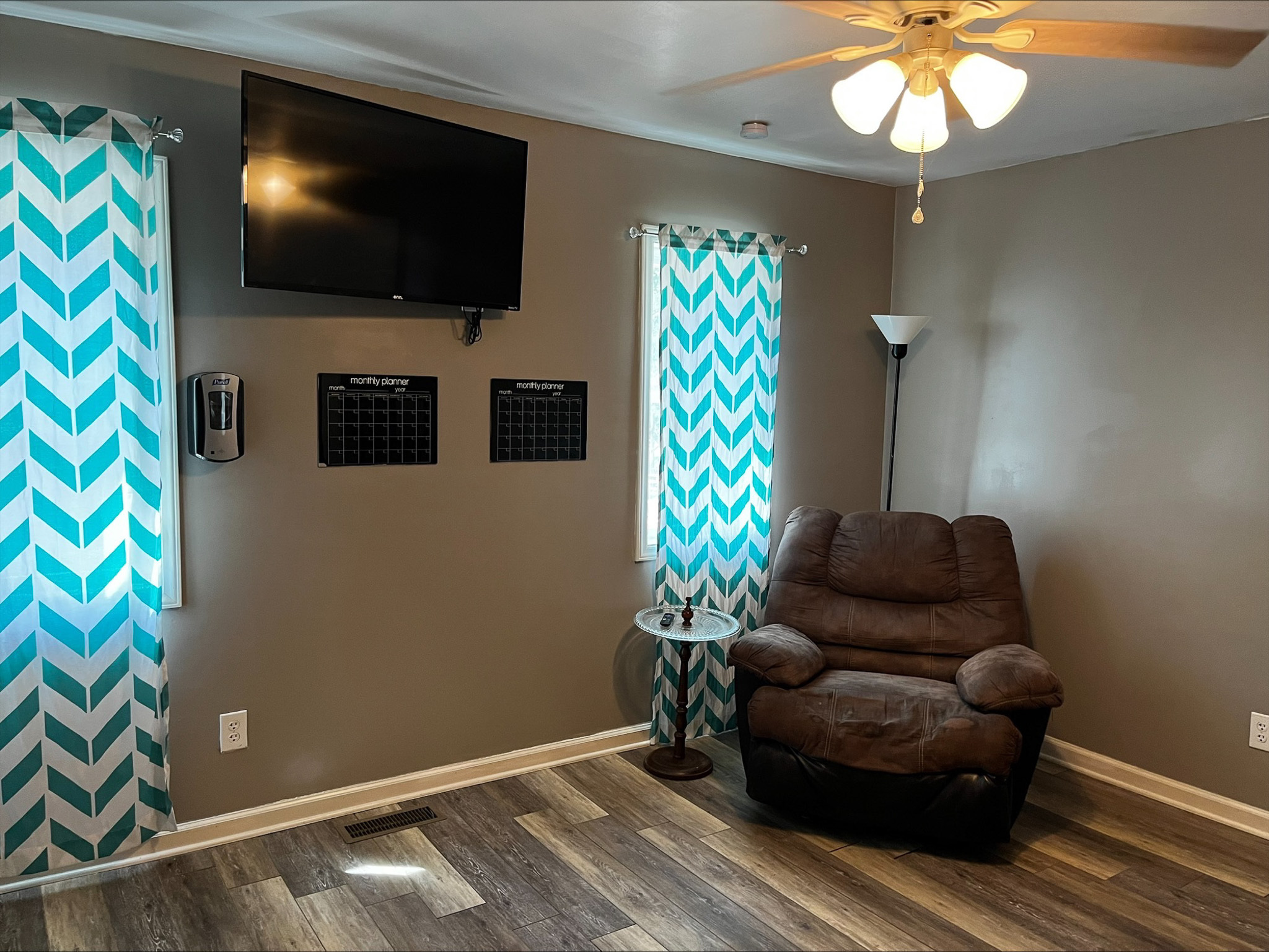Room with gray walls, patterned blue‑and‑white curtains, a mounted TV, brown recliner, and ceiling fan inside the home of Sponsored Residential Provider Dara Short in Willis, Virginia.
