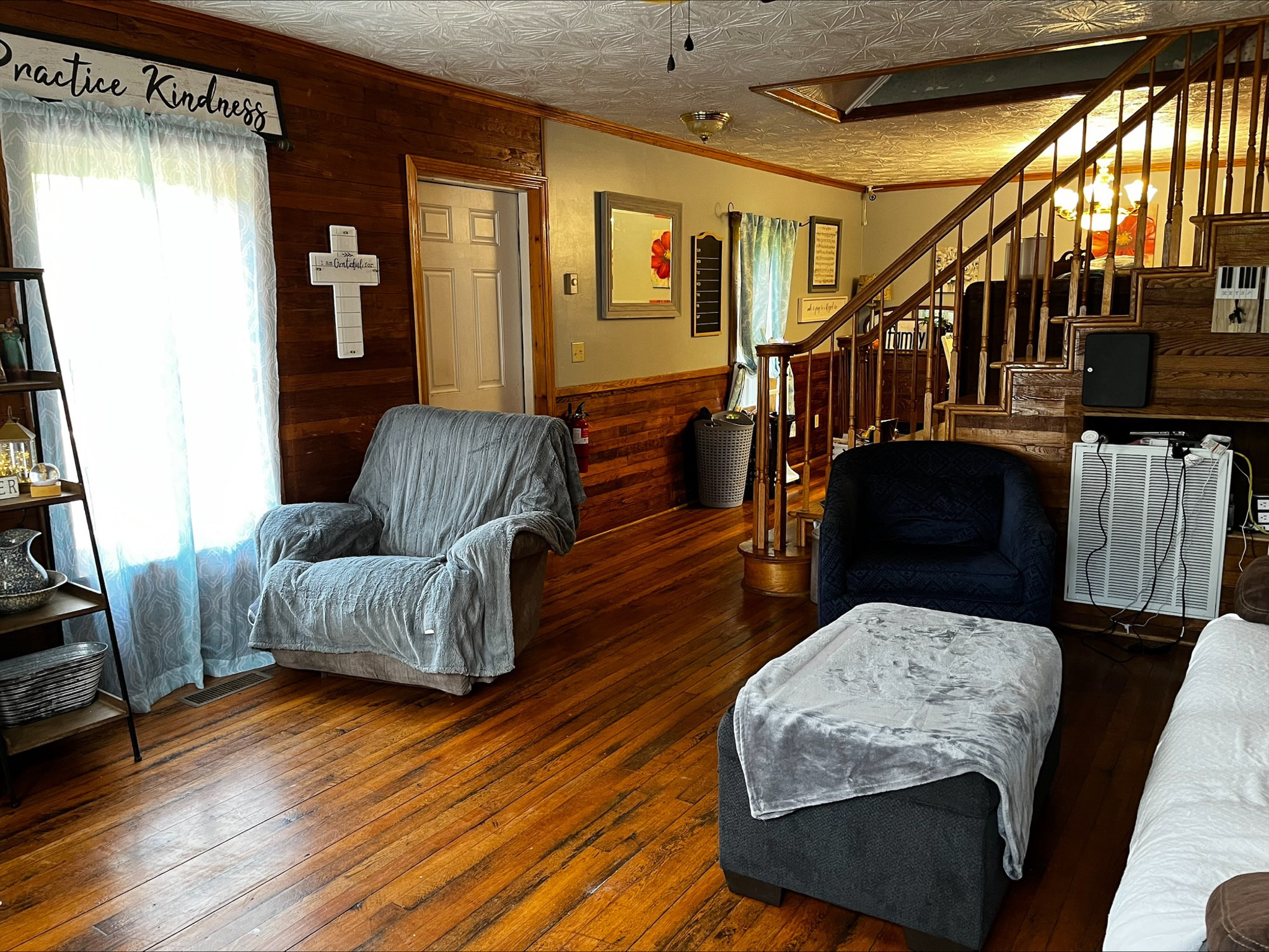 Living room with wood-paneled walls, two armchairs with slipcovers, a large ottoman, hardwood floors, and a staircase with wooden railings in the background inside the home of Sponsored Residential Provider Dara Short in Willis, Virginia.