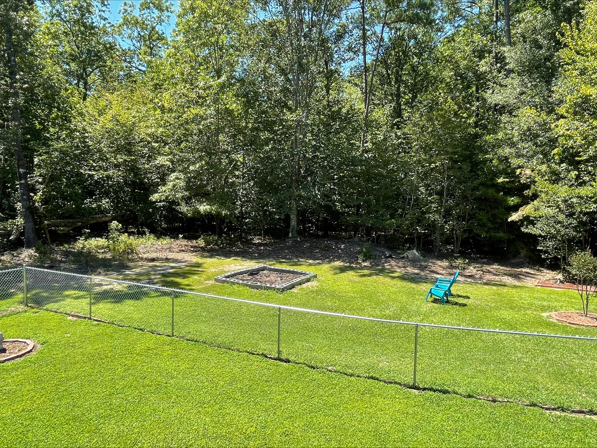 Fenced backyard with a fire pit area, bright green lawn, and dense trees along the back edge, with a single turquoise chair near the fire pit outside the home of Sponsored Residential Provider Kellie Lynch in Gloucester, Virginia.