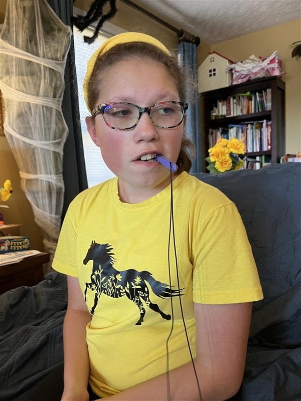 A young girl wearing glasses and a yellow shirt participating in speech therapy, looking toward a speech therapy consultant out of frame